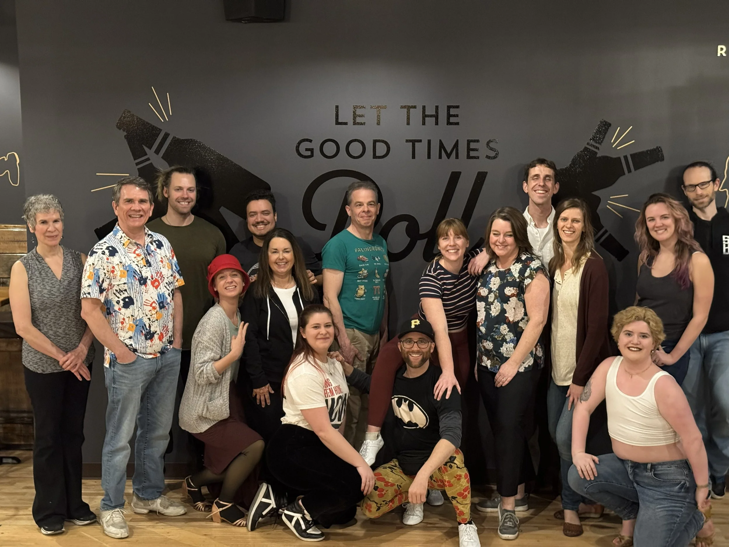 Group of people gathered indoors, smiling for a photo, with a dark wall behind them featuring a large graphic of two bottles and the phrase "Let the Good Times Roll."