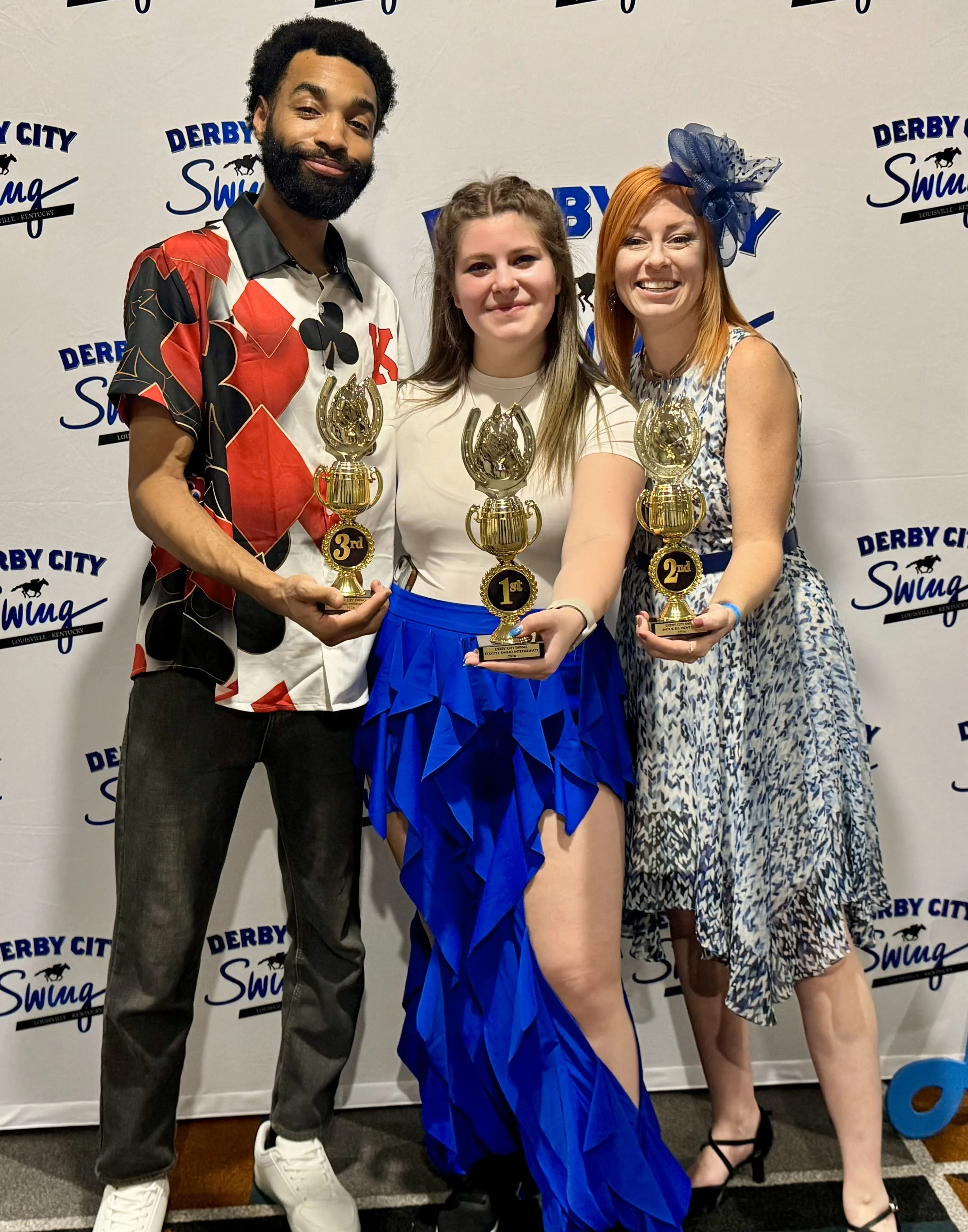 Three people holding trophies, standing in front of a Derby City Swing backdrop. The person in the middle is wearing a white shirt with a blue ruffled skirt, holding a first-place trophy. The person on the left is wearing a colorful patterned shirt, holding a third-place trophy. The person on the right is wearing a patterned dress with a large blue bow headpiece, holding a second-place trophy.