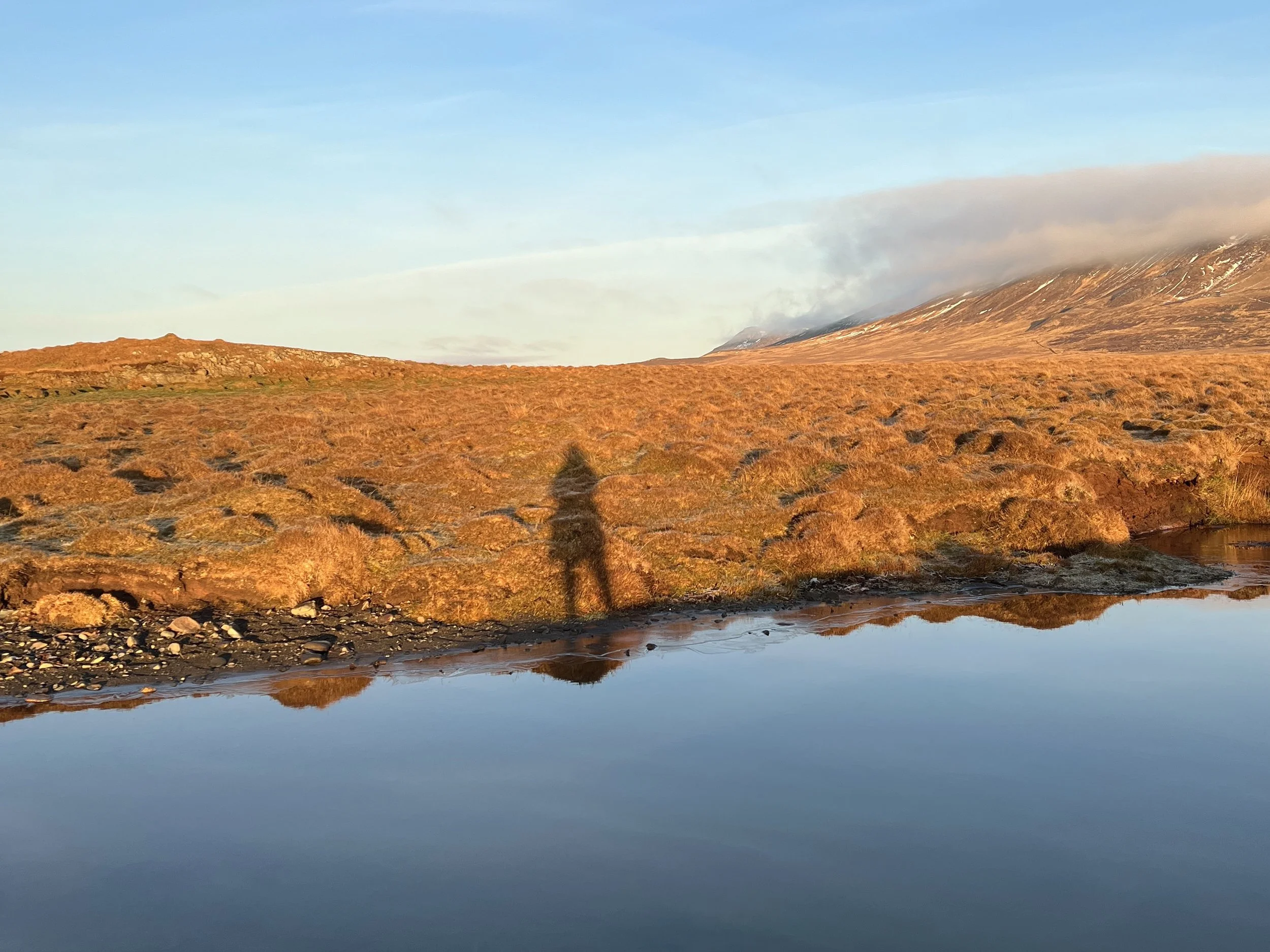 Sunset over a grassy and rocky landscape with a small body of water in the foreground, mountain in the background, and a person's shadow cast on the ground.