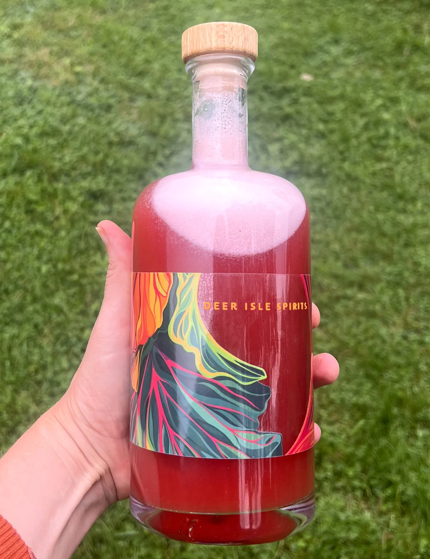 Hand holding a clear glass bottle with a wooden cap, decorated with colorful abstract artwork, and the words 'Deer Isle Spirits,' filled with a pinkish-red beverage, outdoors on grass.