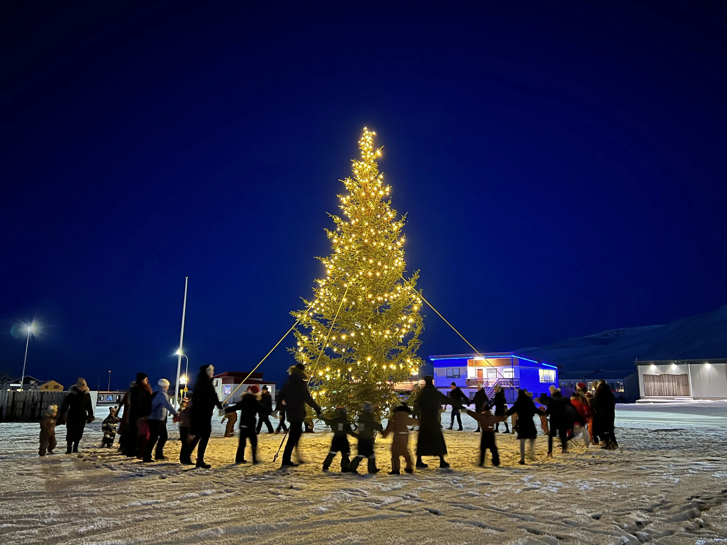 People holding hands in a circle around a decorated Christmas tree with yellow lights during nighttime in a snowy outdoor setting.