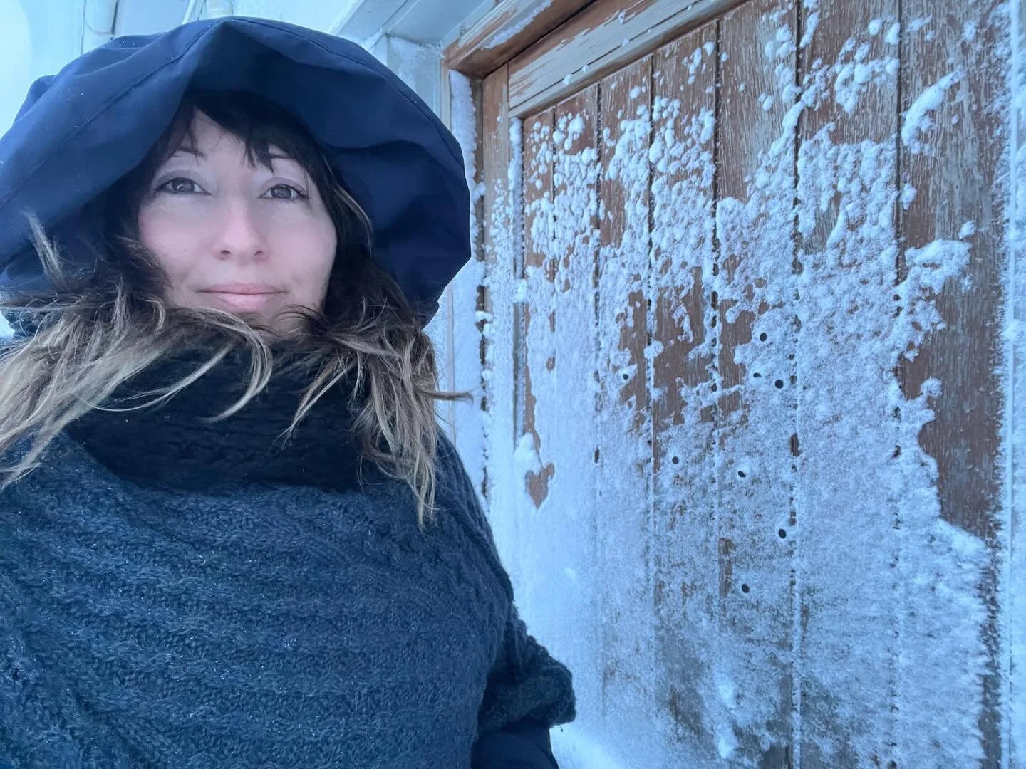 A woman with long, wavy hair, wearing a dark hooded jacket, stands outdoors in front of a snow-covered wooden door. She is looking at the camera with a slight smile, and snow is visible on her jacket and the door.