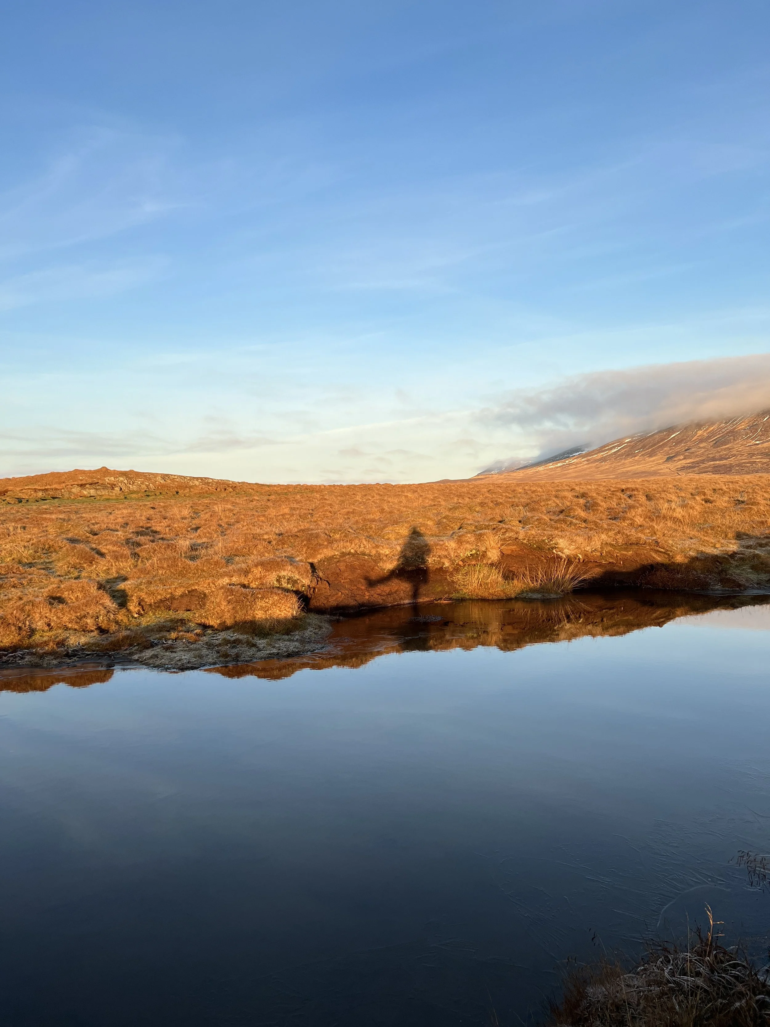 A landscape with a calm water body reflecting the sky, dry grass, and a mountain in the background, all under a blue sky with clouds.