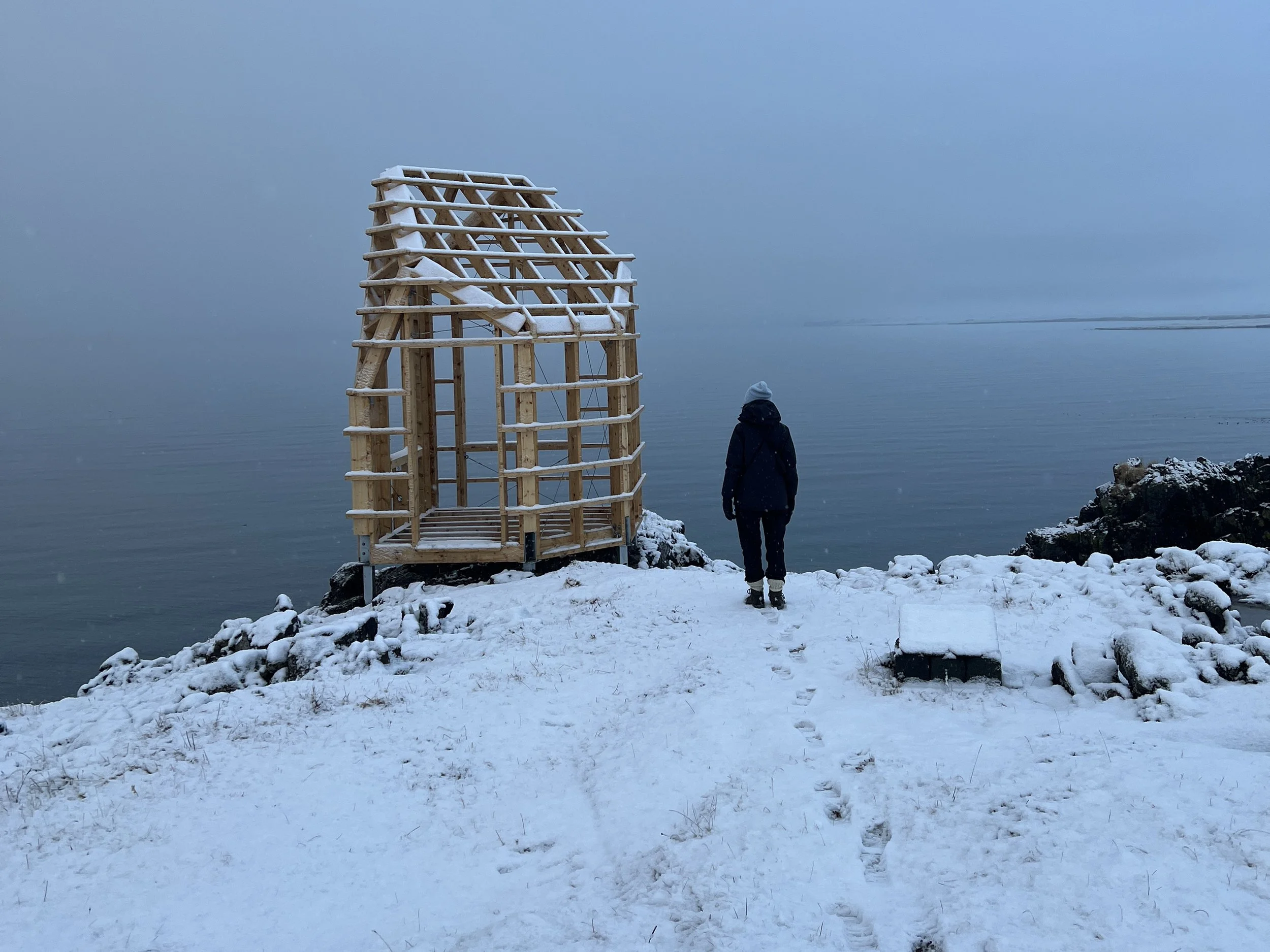 A person dressed in winter clothing walking towards a small wooden structure by a snowy shoreline, with a calm body of water and overcast sky in the background.