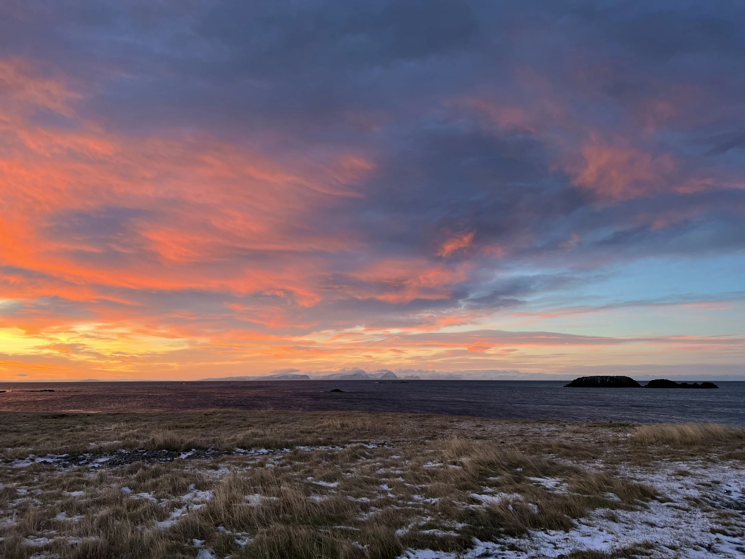 Sunset over the ocean with pink, orange, and blue clouds, grassy shoreline with patches of snow, and small islands in the distance.