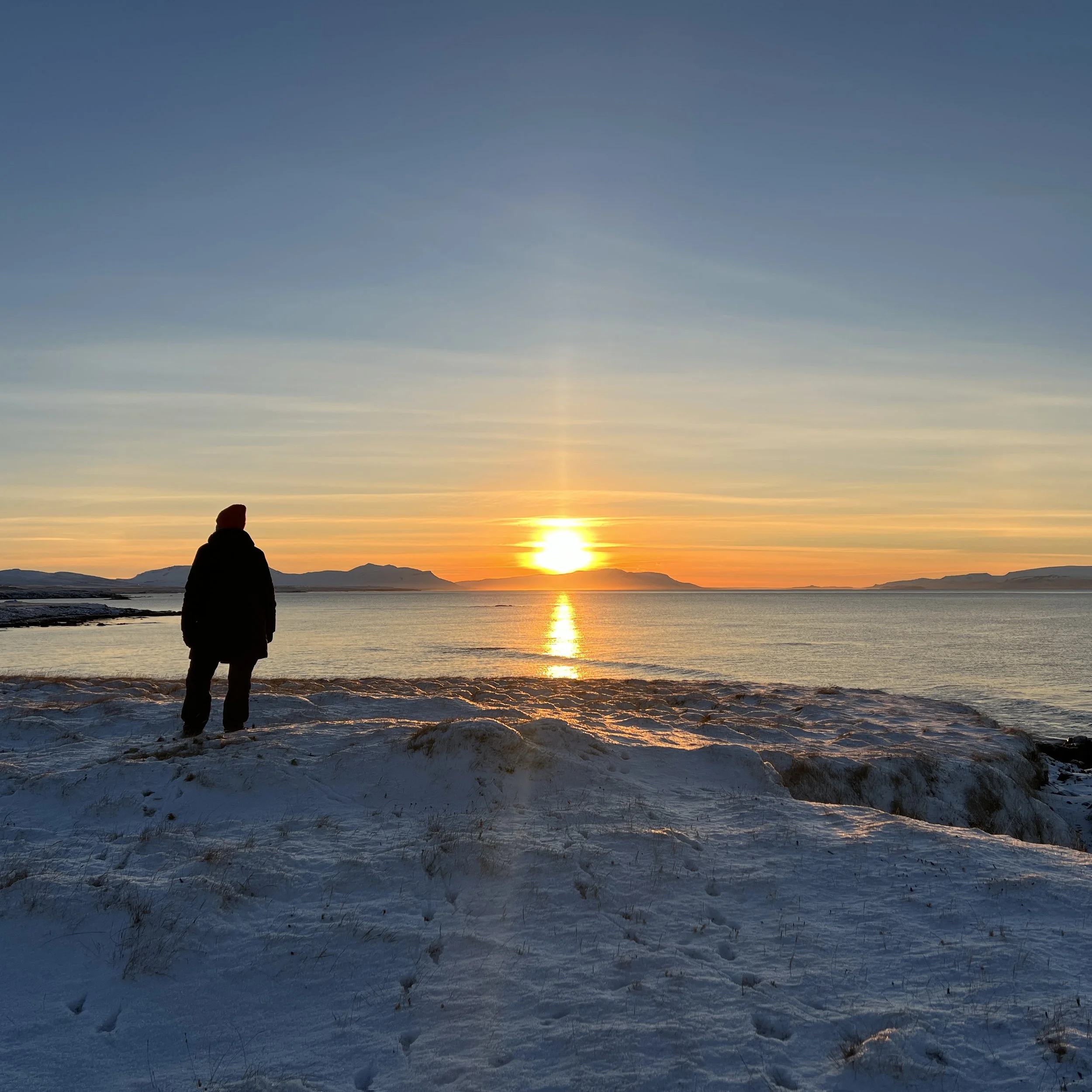 Person standing on snowy shore watching a sunset over a calm body of water with mountains in the distance.