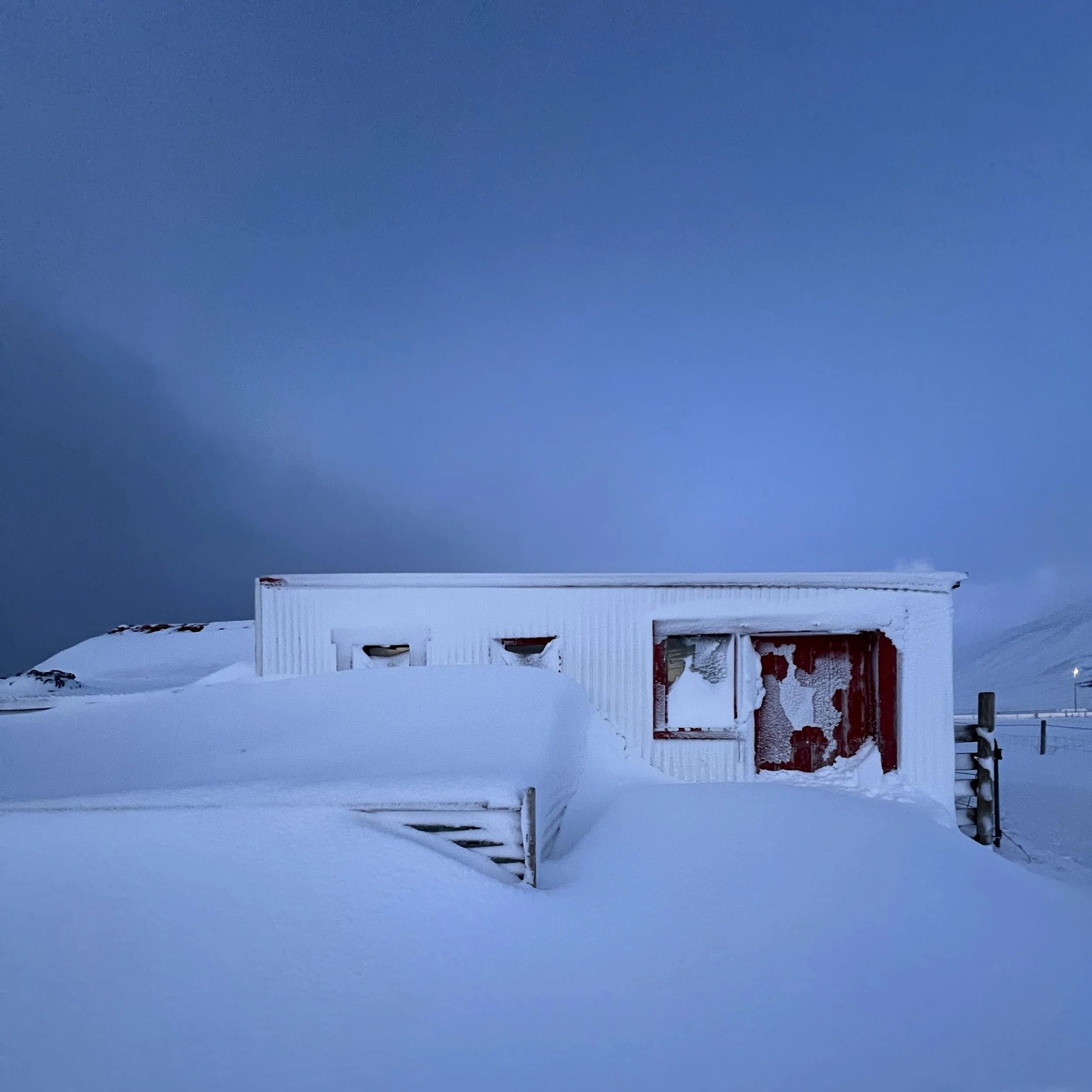 A small building surrounded by deep snow, with a partially snow-covered staircase and a large snowbank in front. The building has a white exterior with a broken window, against a dark, overcast sky.
