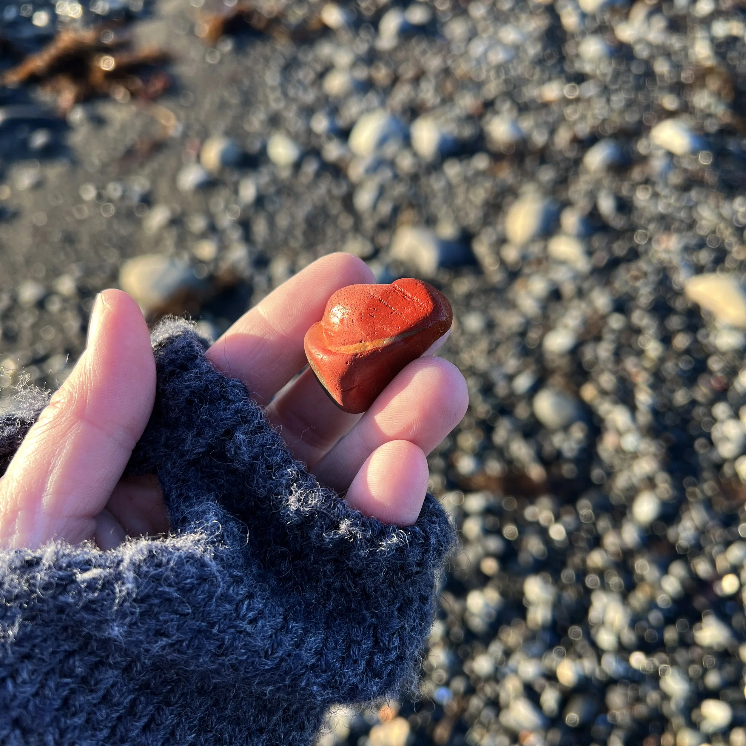 Person holding a polished red heart-shaped stone in their hand, wearing fingerless gray gloves, with a rocky beach background.