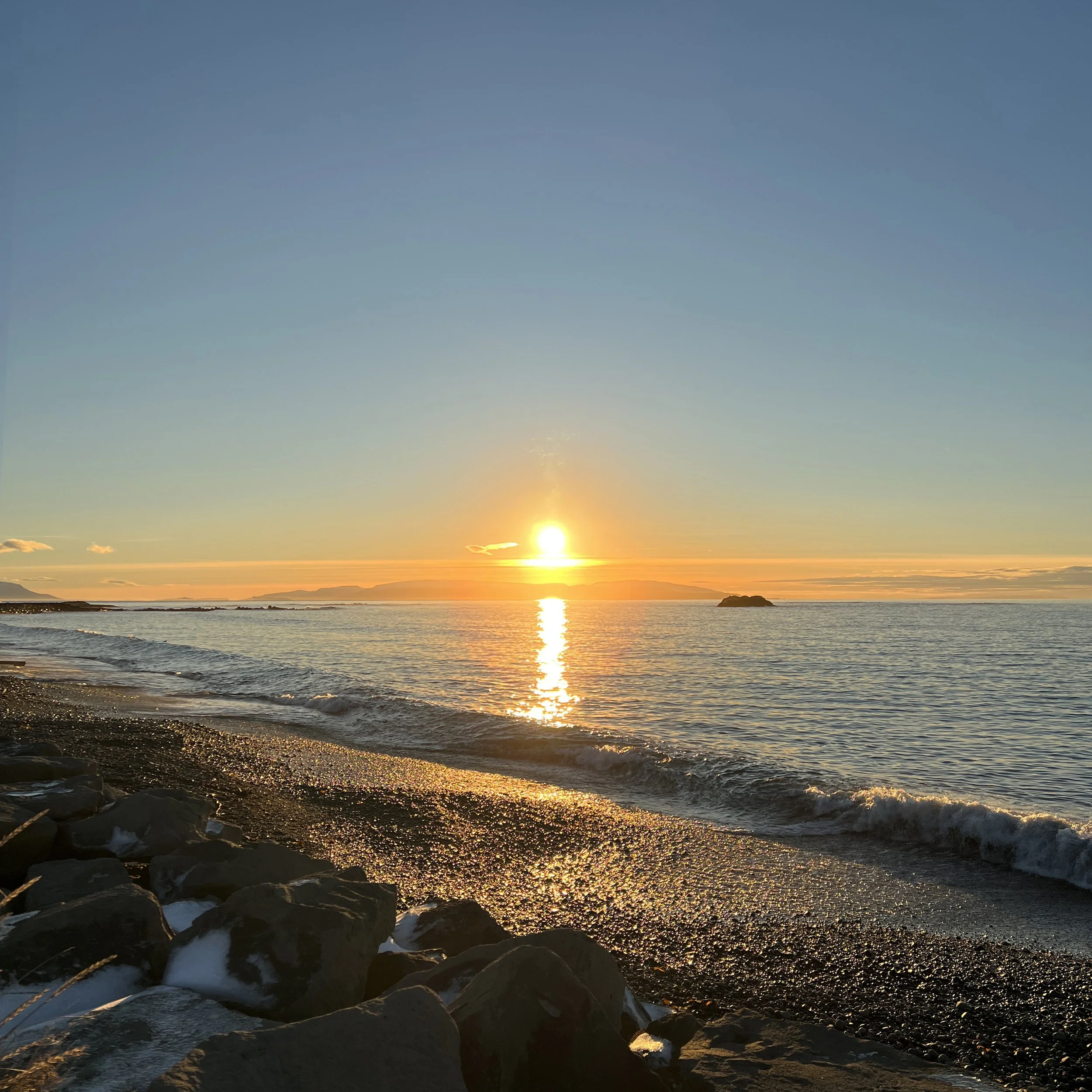 Sunset over the ocean with calm waves hitting the rocky shoreline, reflected light from the sun shimmering on the water, and distant land in the background.