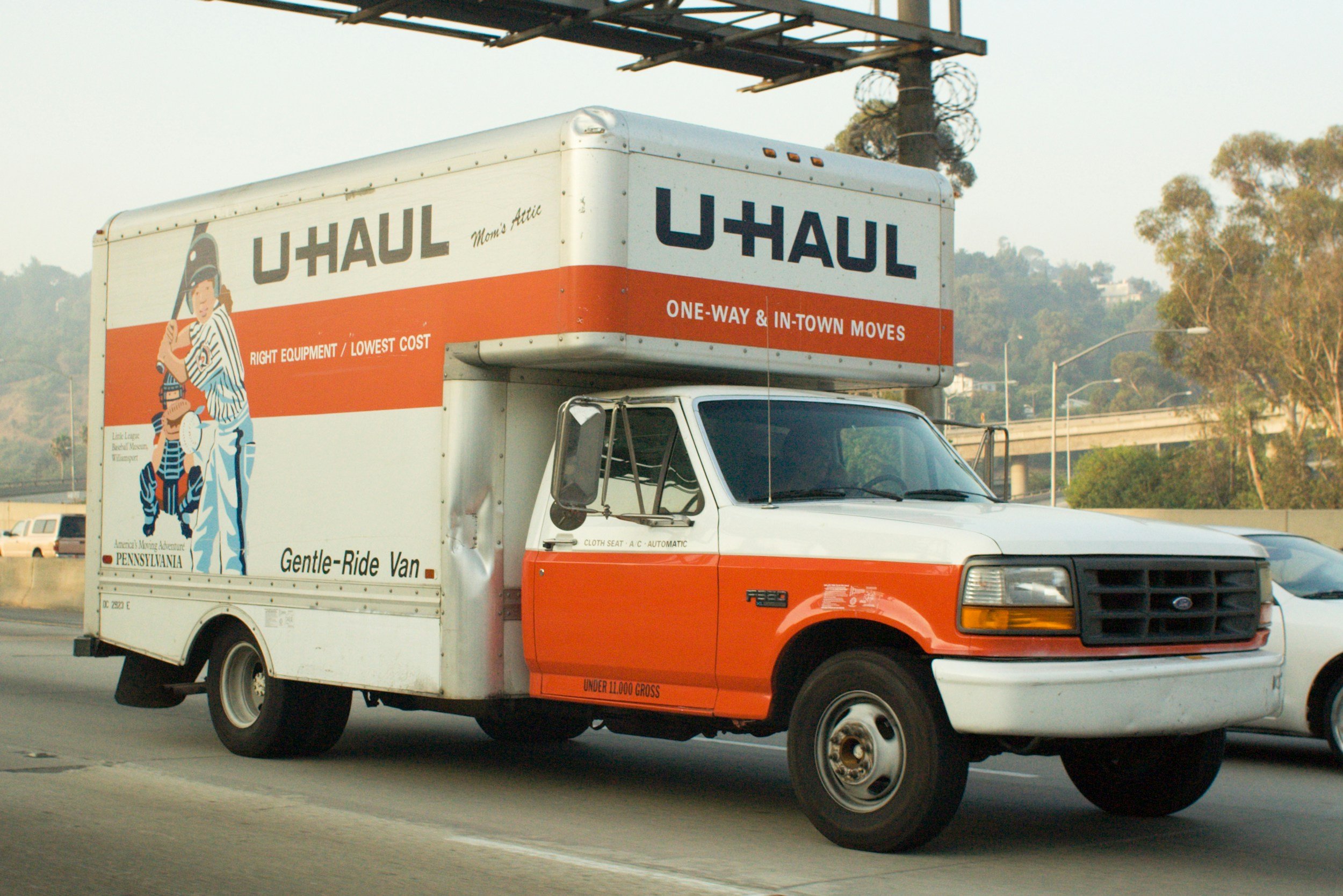 A U-Haul truck driving on a highway, with a white cab and orange cargo area, featuring illustrations and text about moving services.