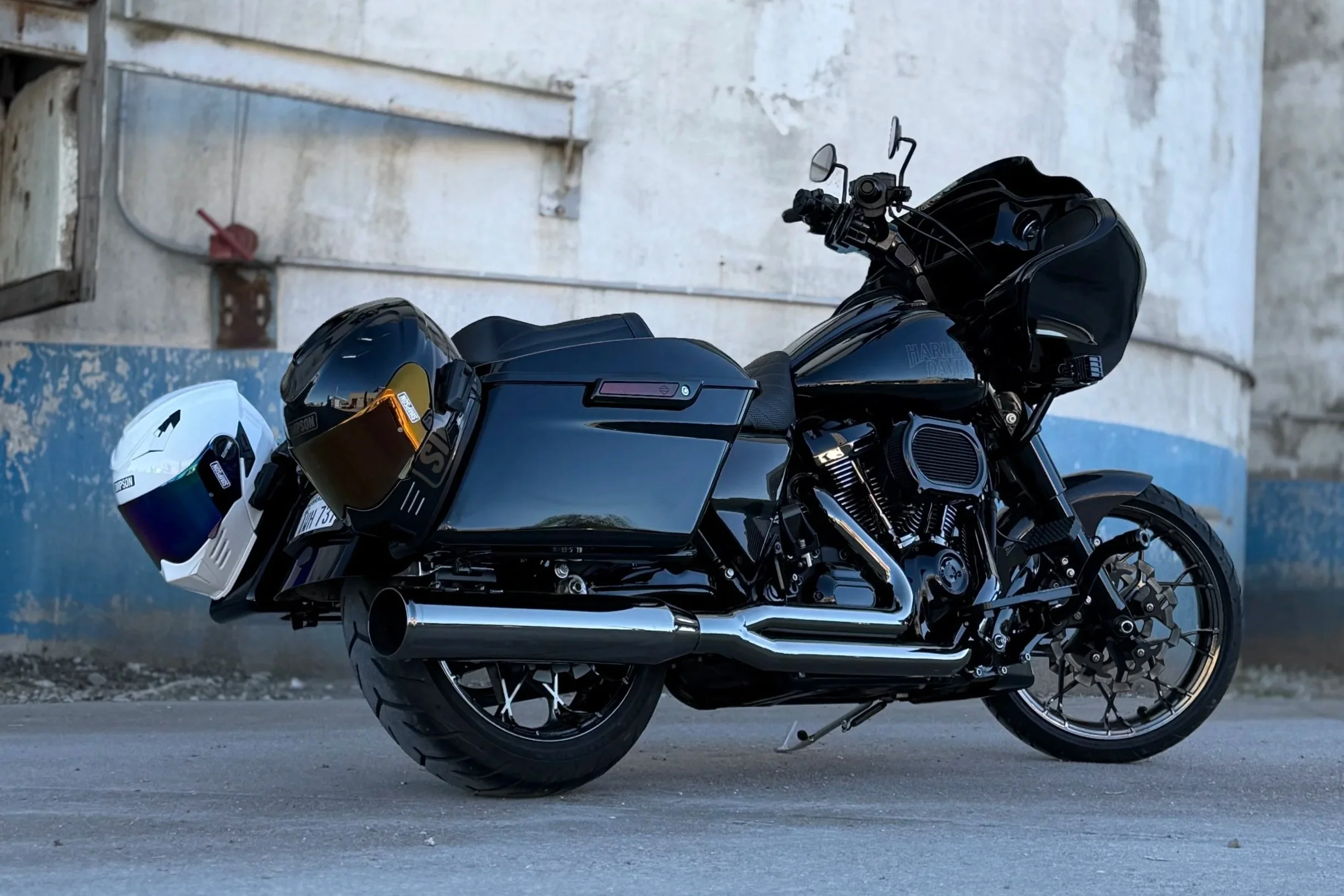 Black Harley Davidson motorcycle with two helmets, one white and the other black with gold visor, parked near a weathered wall.