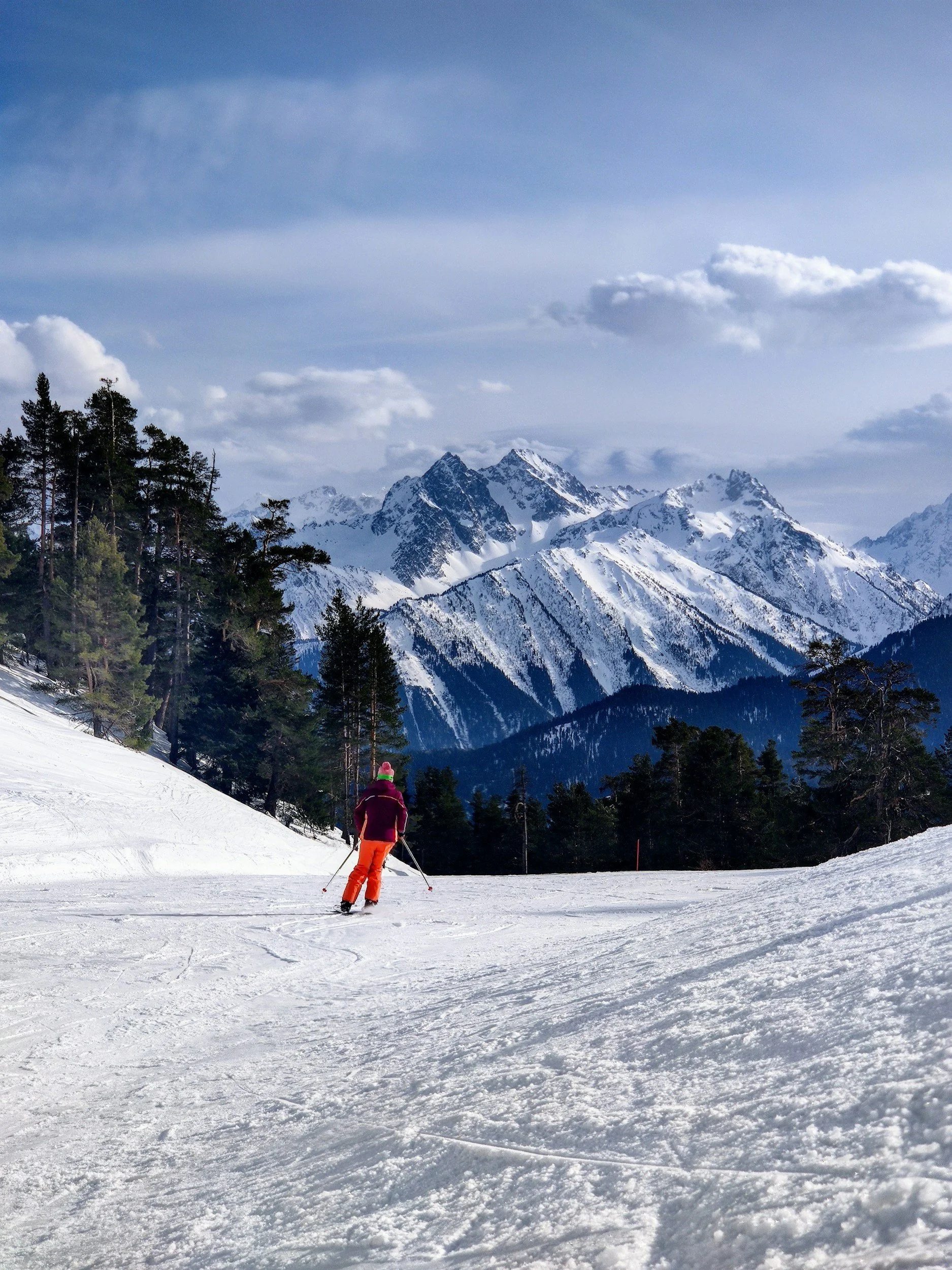A person in pink and orange winter clothing skiing on a snowy mountain slope with tall pine trees and snow-covered mountains in the background under a blue sky with scattered clouds.