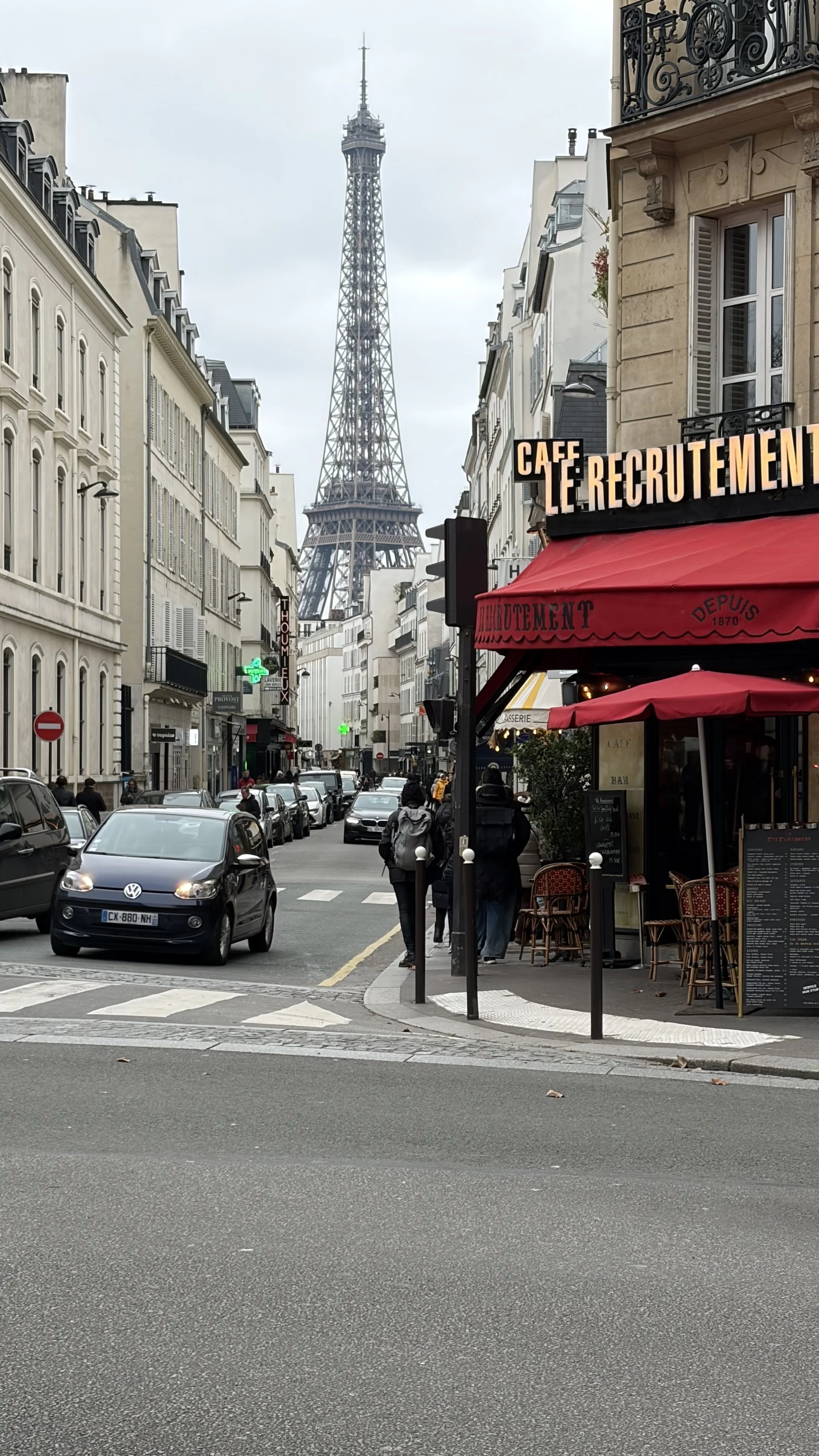 A street scene on a cloudy day in Paris with the Eiffel Tower in the background. Cars are parked along the street, and pedestrians are walking on the sidewalk. There is a café with outdoor seating and a sign that reads 'CAFÉ RECRUTEMENT' on a red awning.