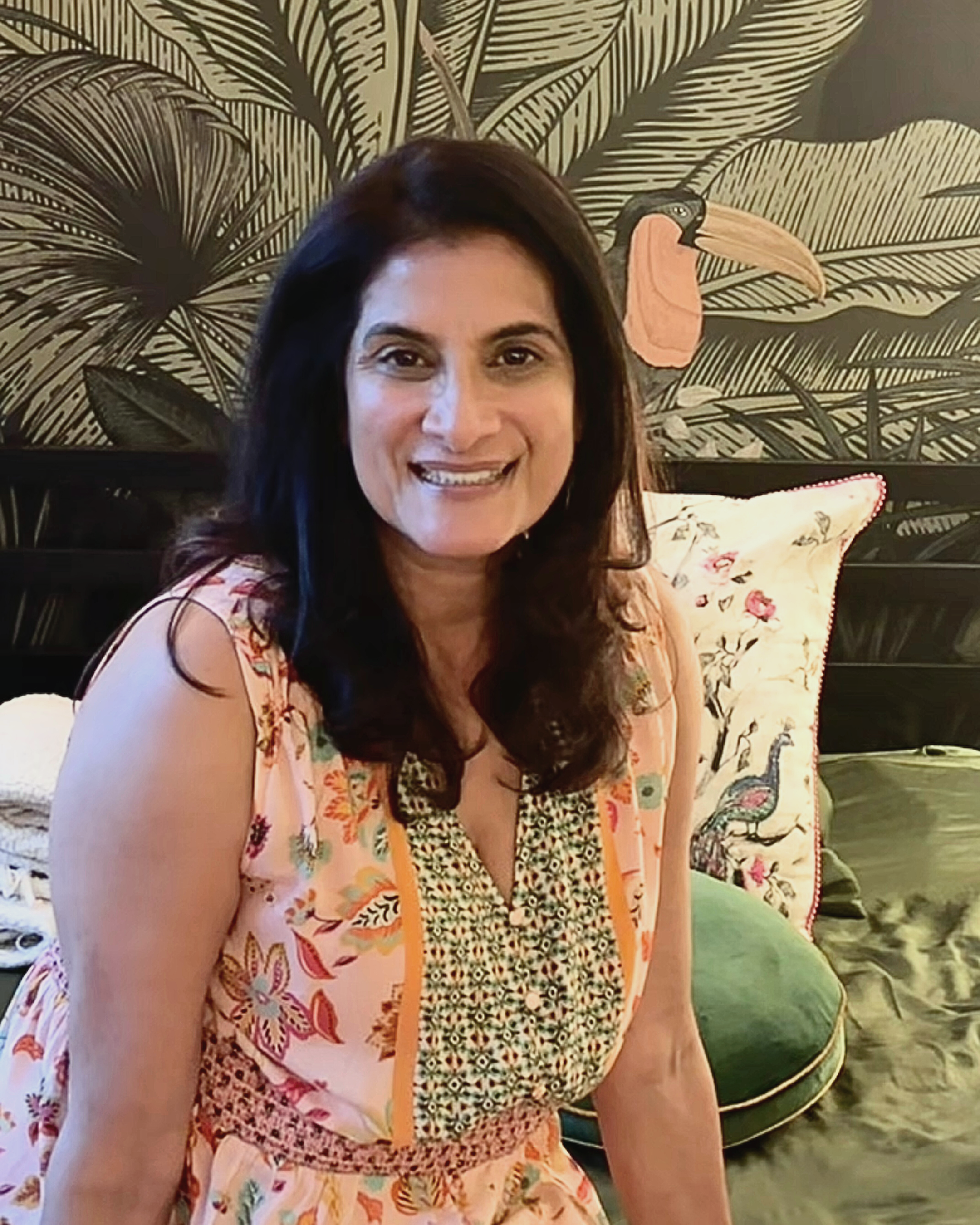 Anjali Augustine with dark brown hair and a floral sleeveless dress sitting on a floral-covered chair in front of a black mural with large leaves and a toucan painting in the background.