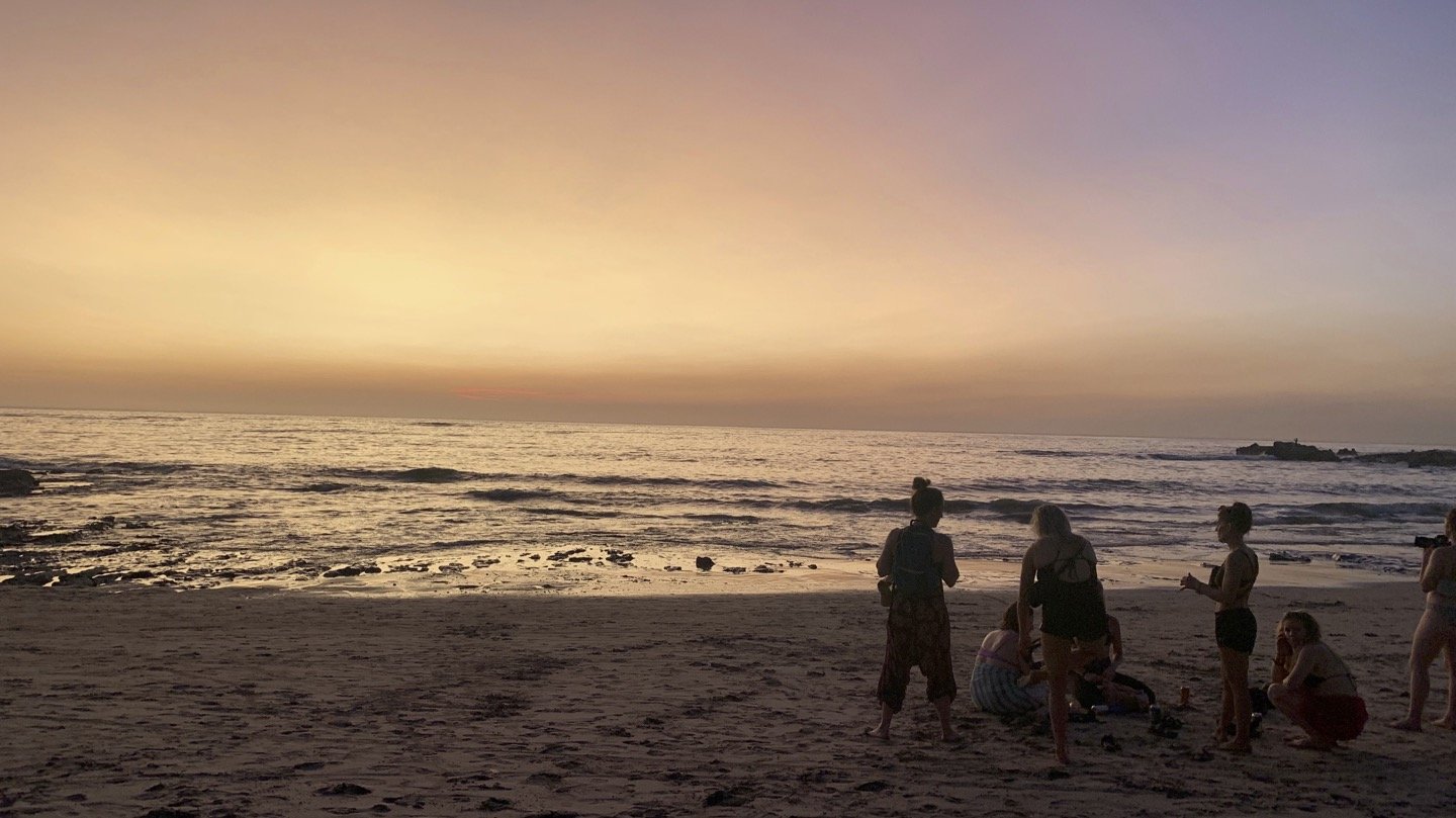 People gathered on a beach during sunset, with a colorful sky and calm ocean waves.