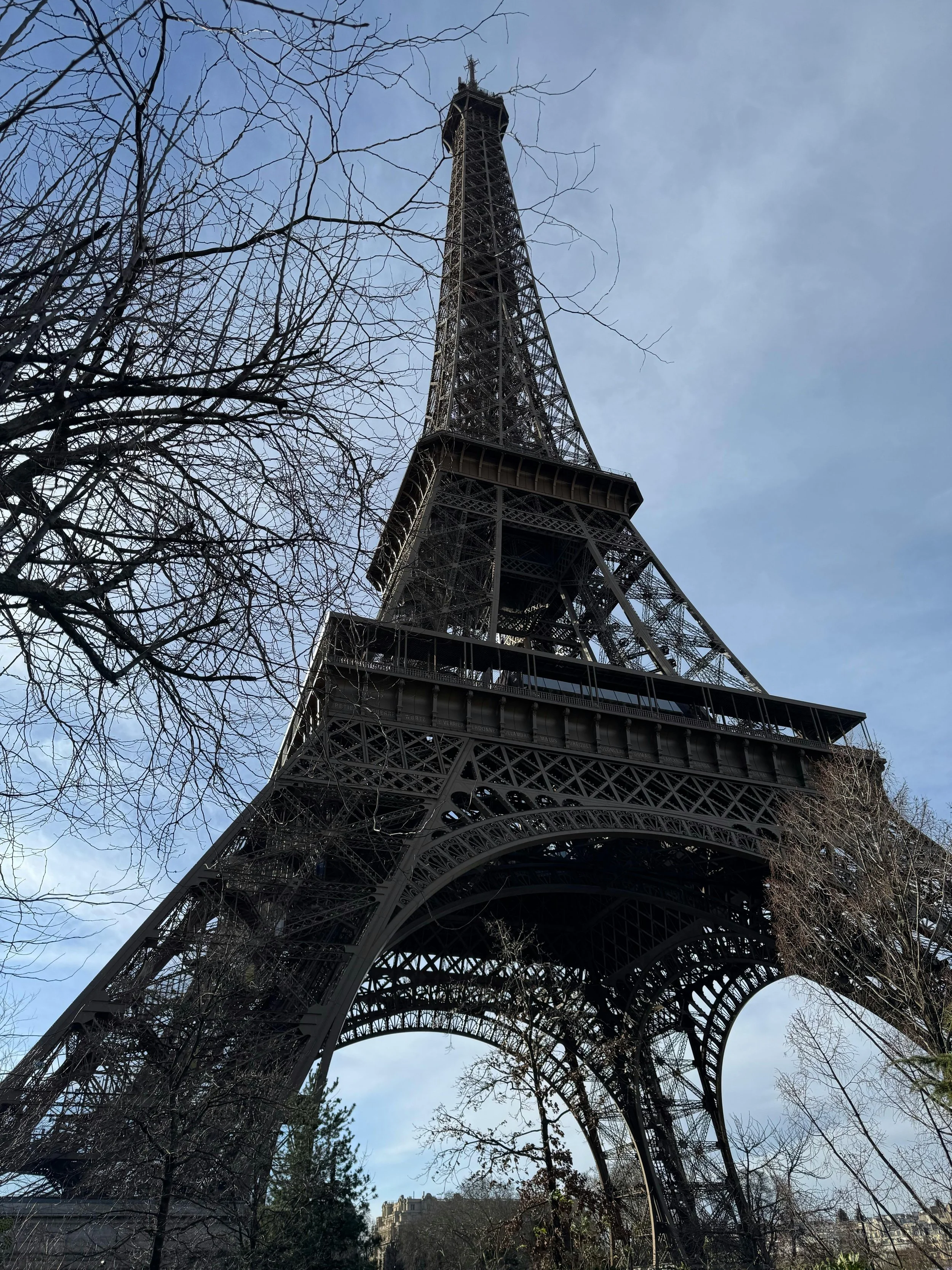 Eiffel Tower in Paris viewed from below with a partly cloudy sky and leafless trees in the foreground.