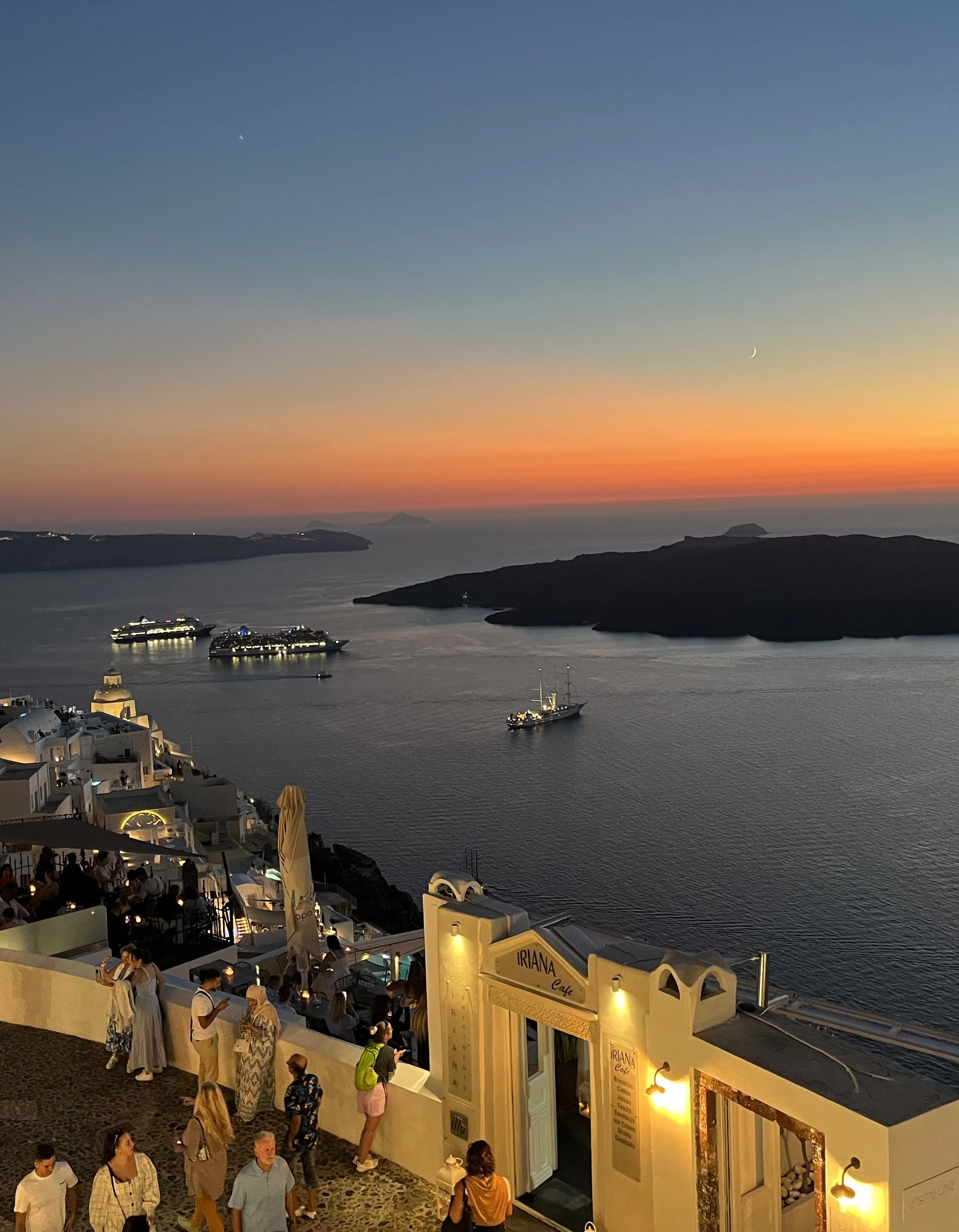 People gathered on a terrace overlooking the ocean at sunset in Santorini, Greece, with cruise ships on the water and islands in the background.