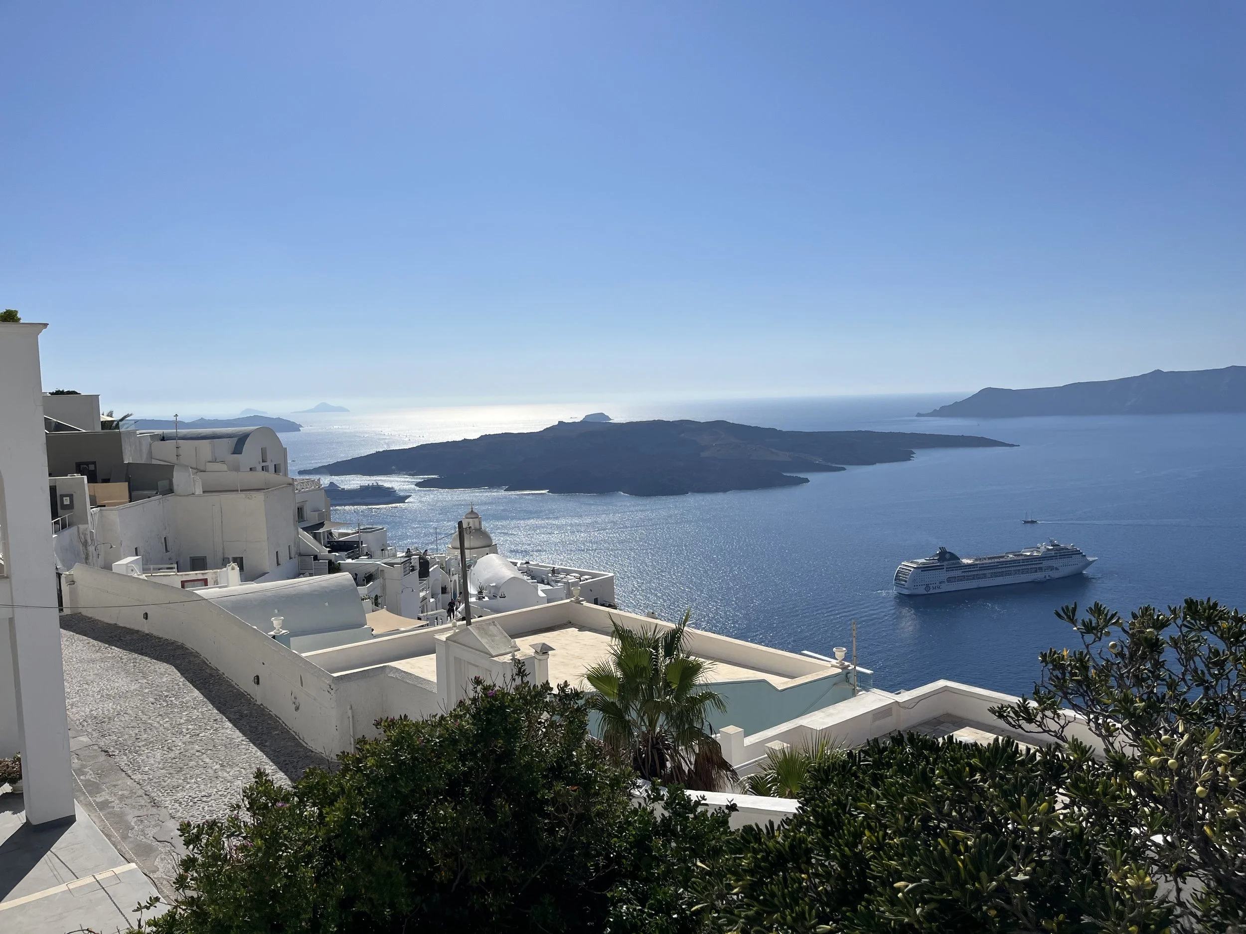 White buildings with domed roofs on the island of Santorini in Greece on a hillside overlooking the sea, with mountains and a cruise ship in the water under a clear blue sky.