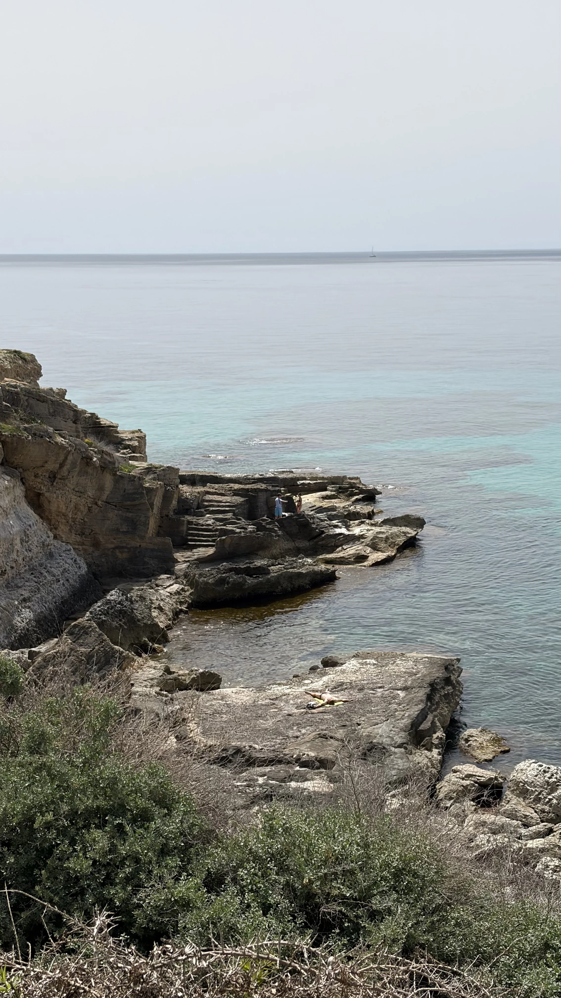 Rocky shoreline in Mallorca with a few people sitting and standing on the rocks, overlooking calm ocean water, with a sailboat in the distance and a cloudy sky.