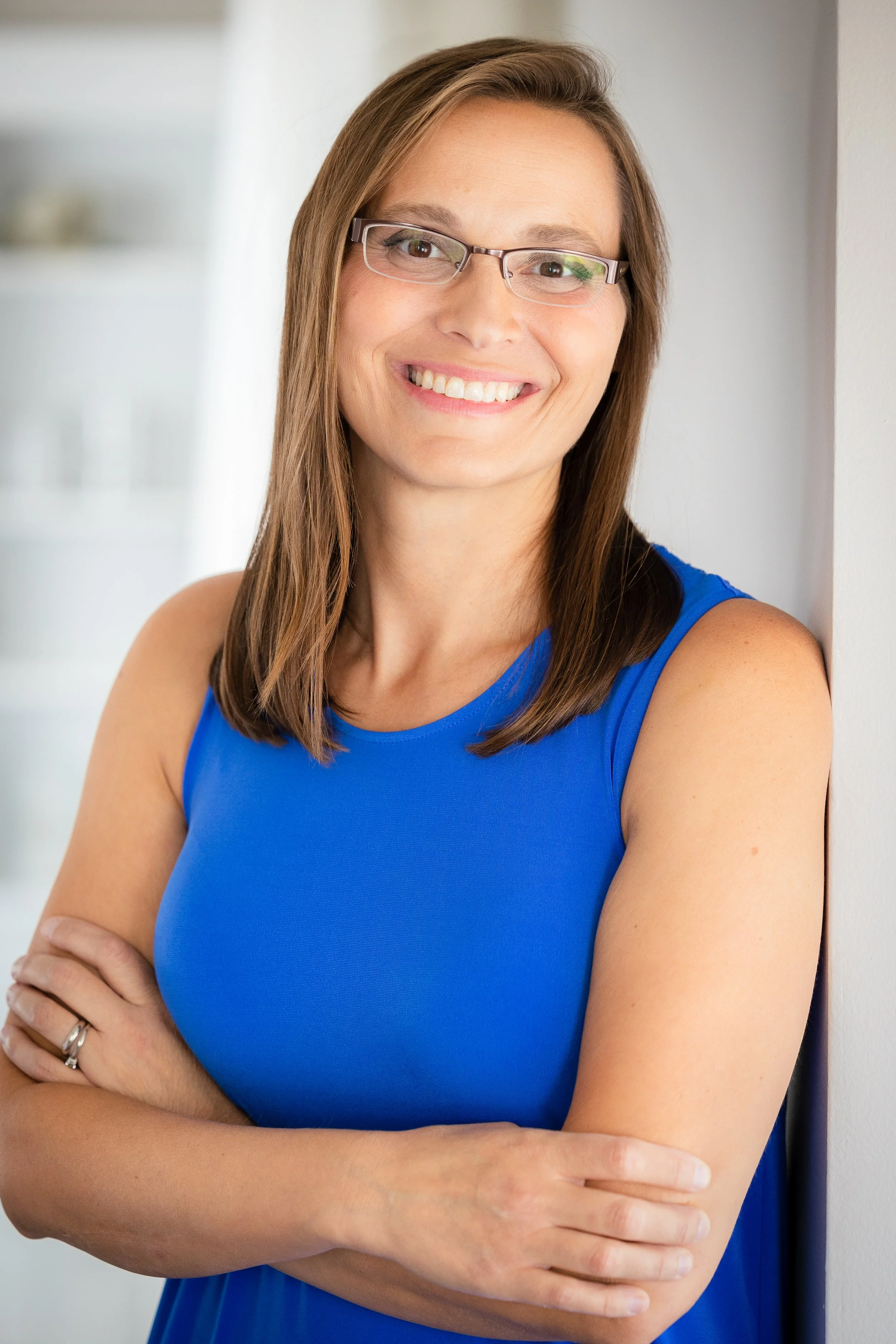 A woman with glasses and shoulder-length brown hair, smiling, standing with arms crossed against a white wall, wearing a sleeveless blue top.