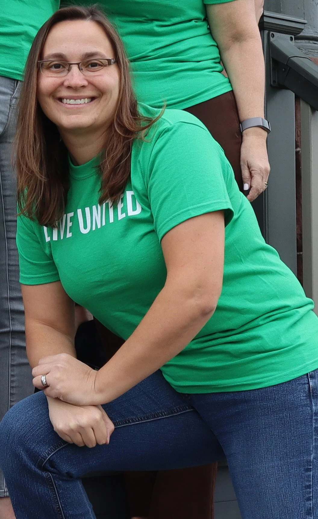 A woman with brown hair, glasses, and a green shirt kneeling with her hands clasped resting on her leg.