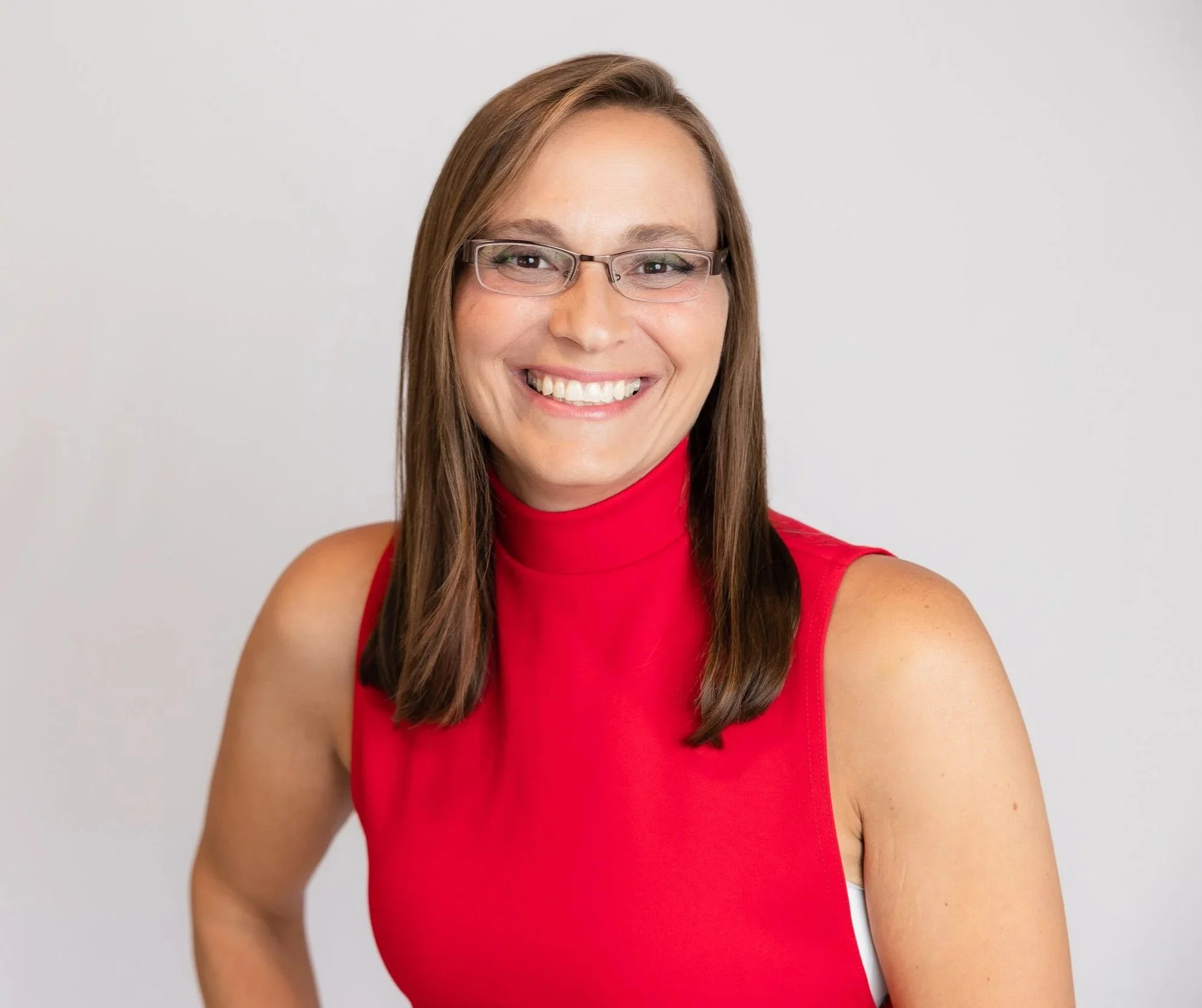 A woman smiling, wearing glasses and a sleeveless red top, with straight brown hair, against a neutral background.
