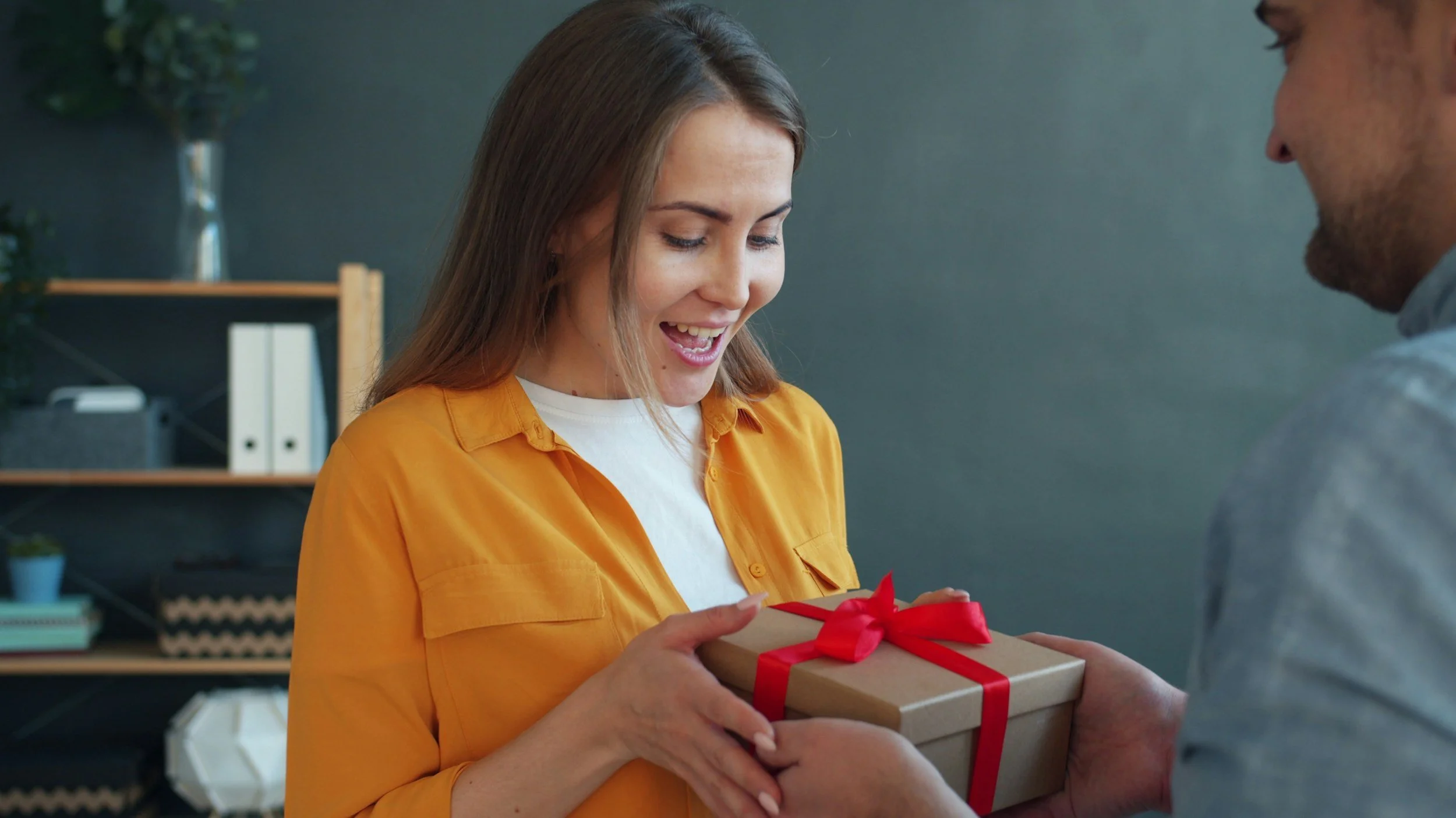 A woman in a yellow shirt is smiling as she receives a gift wrapped with a red ribbon from a man who is partially visible. They appear to be indoors with a bookshelf and decorations in the background.