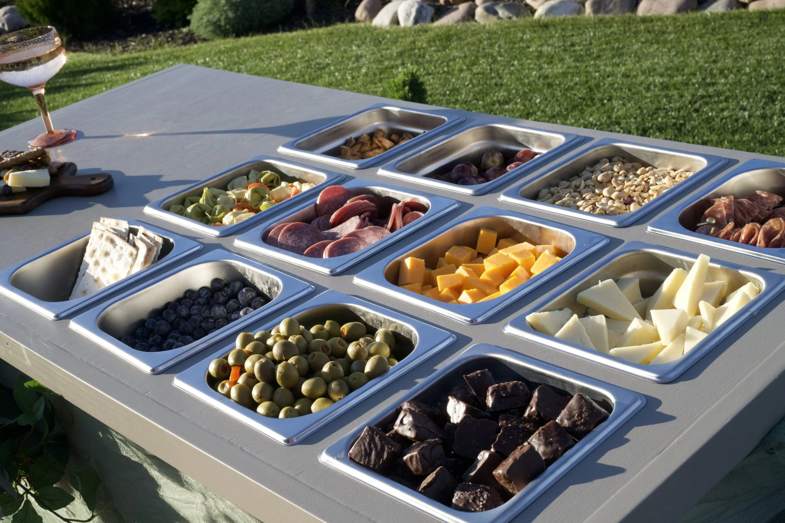 A white table outdoors with various cheeses, meats, olives, grapes, nuts, and chocolates arranged in metal trays for a charcuterie spread.