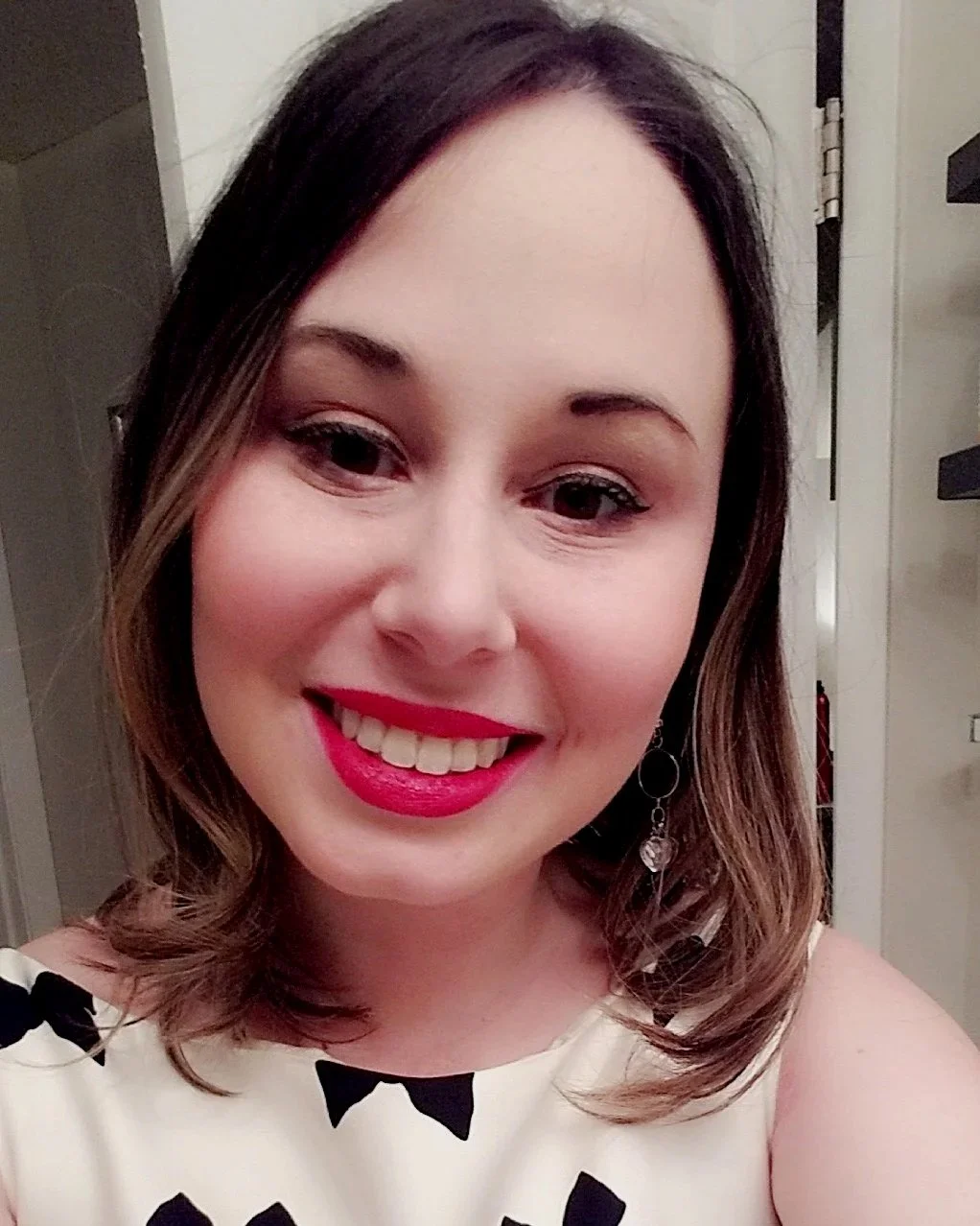 A woman with shoulder-length brown hair, wearing bright red lipstick, smiling, and dressed in a white top with black bow patterns.