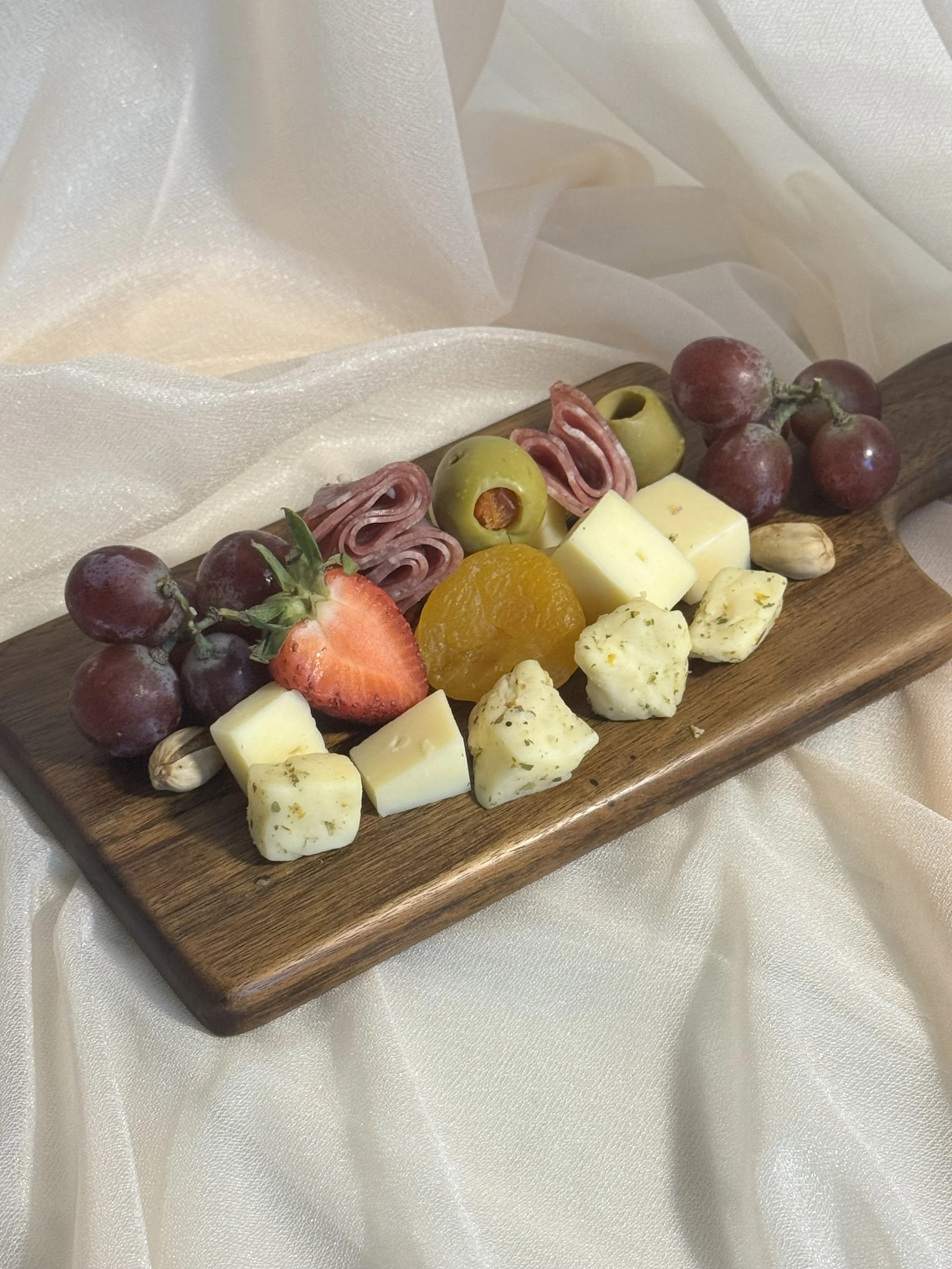 A wooden serving board with assorted cheese, grape, strawberry, dried apricot, green olives, ham rolled slices, garlic, and cheese chunks, set on cream-colored satin fabric.