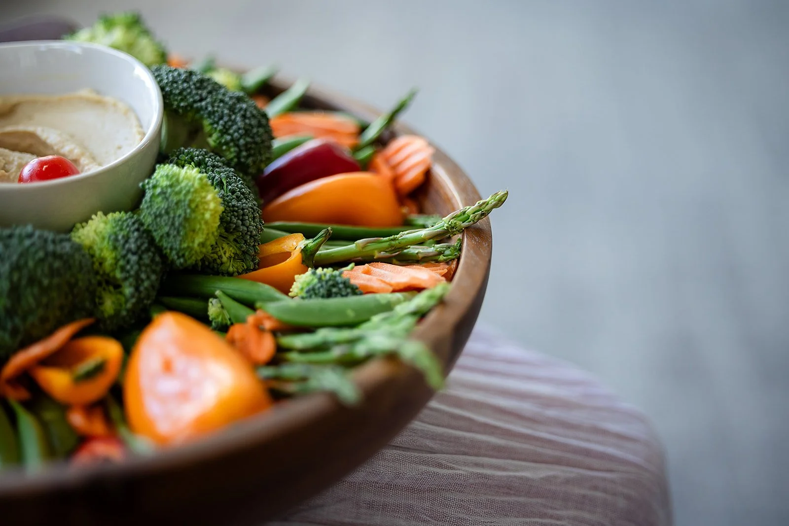 A round wooden platter filled with broccoli florets, cherry tomatoes, orange and yellow bell pepper slices, green beans, and asparagus, with a small cup of dip or dressing on the side.