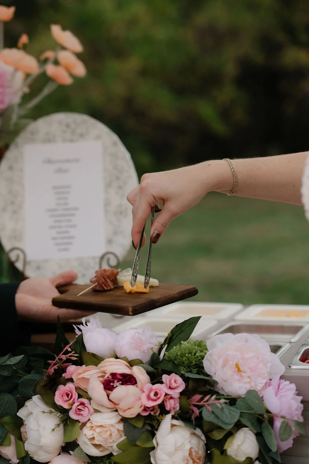 Person serving cheese and meat to another person at a wedding or celebration, with pink and white flowers in the foreground and a blurred menu in the background.