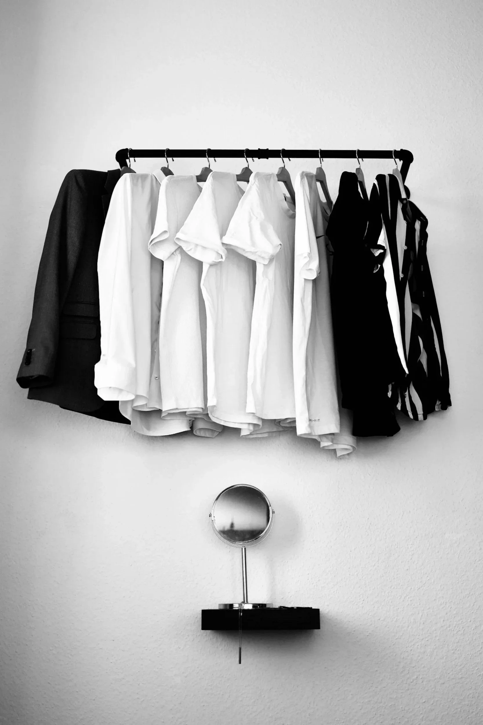 Black-and-white photo of a clothing rack with white, black, and striped shirts, and a black blazer, with a small round mirror below.