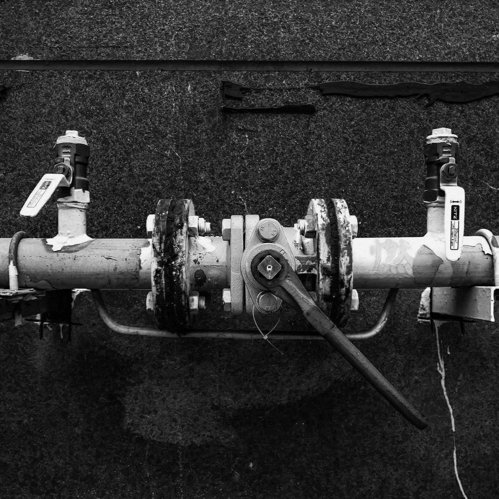 Black and white photo of a metal pipe with valves and a lever, mounted on a textured dark surface.