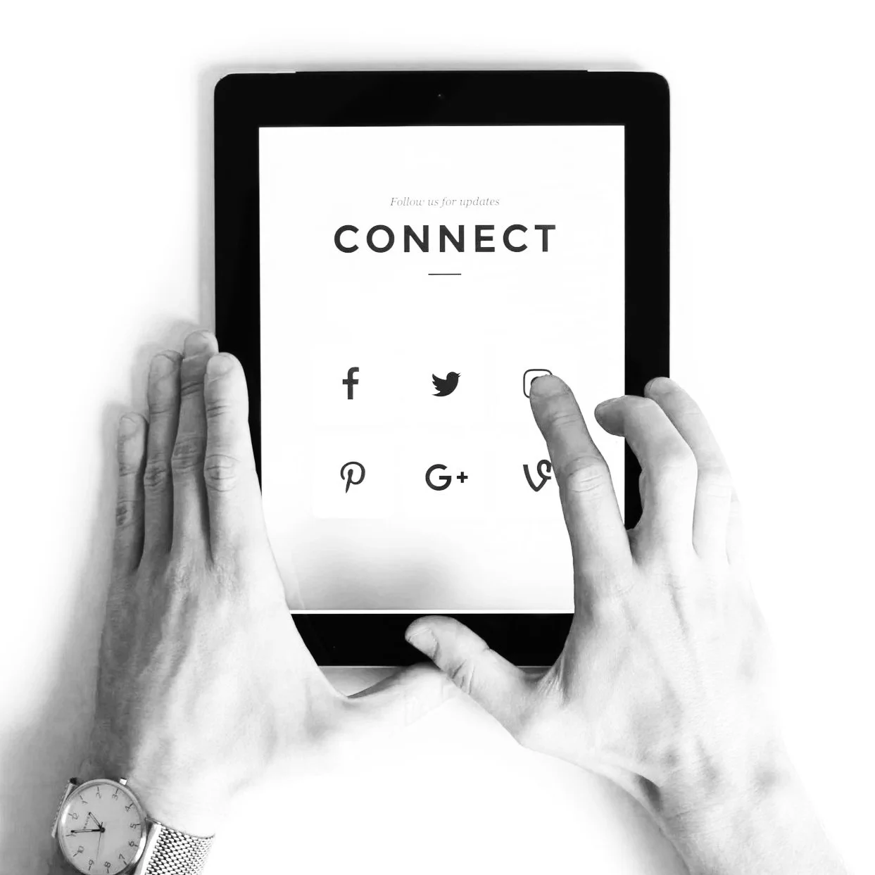 Black and white photo of a person holding a tablet showing social media icons, with hands visible and wearing a watch on the left wrist.
