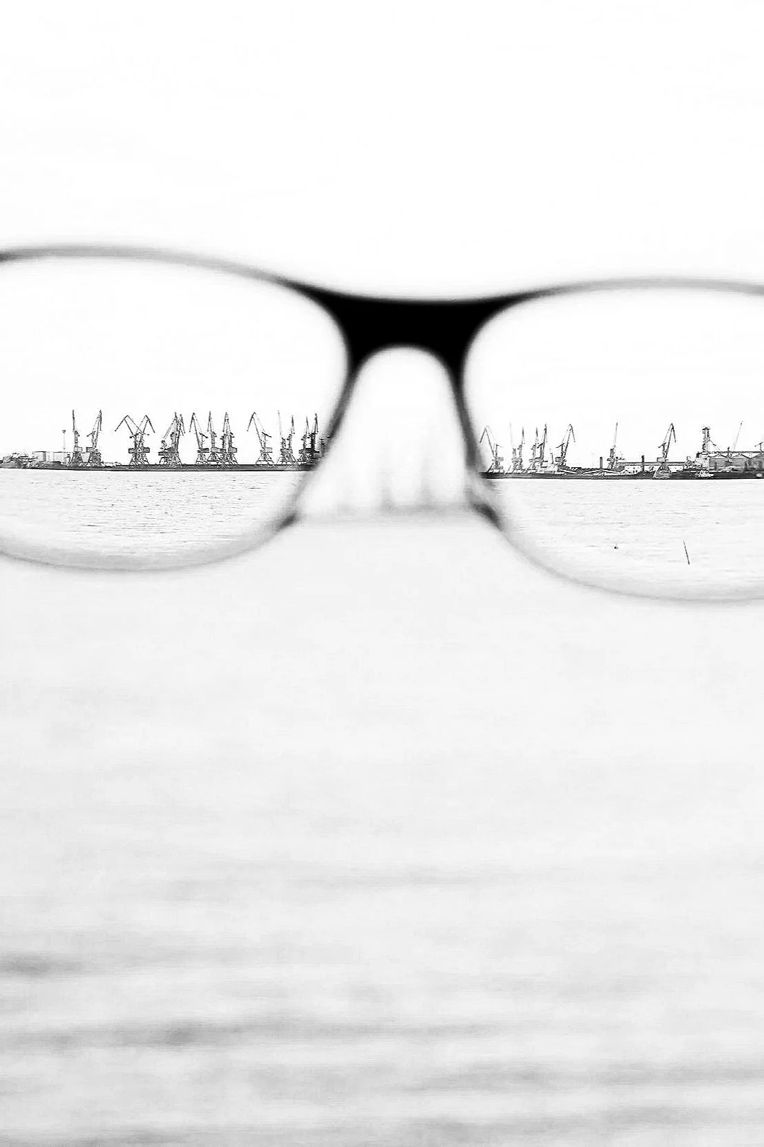 Close-up of glasses with a harbor and cranes visible through the lenses in black and white.