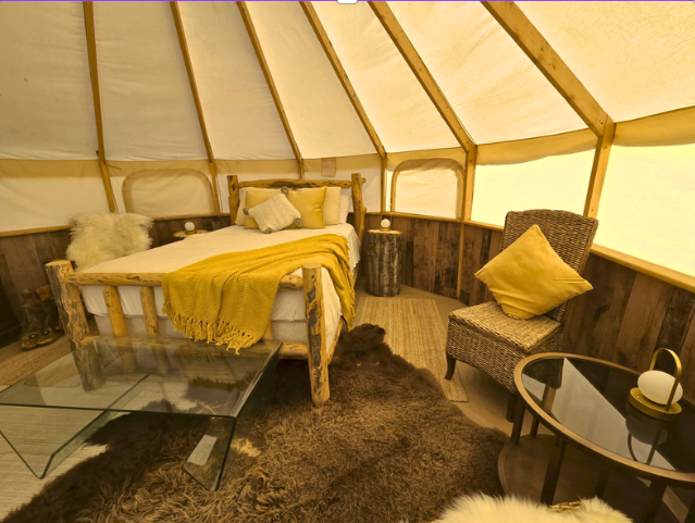 Interior view of a cozy, rustic bell tent bedroom with a post and beam bed, yellow blanket, pillows, wicker chair with a yellow pillow, glass coffee table, small round side table, surrounded by animal fur rugs.  TK Glamping and Wellness in Vernon BC.
