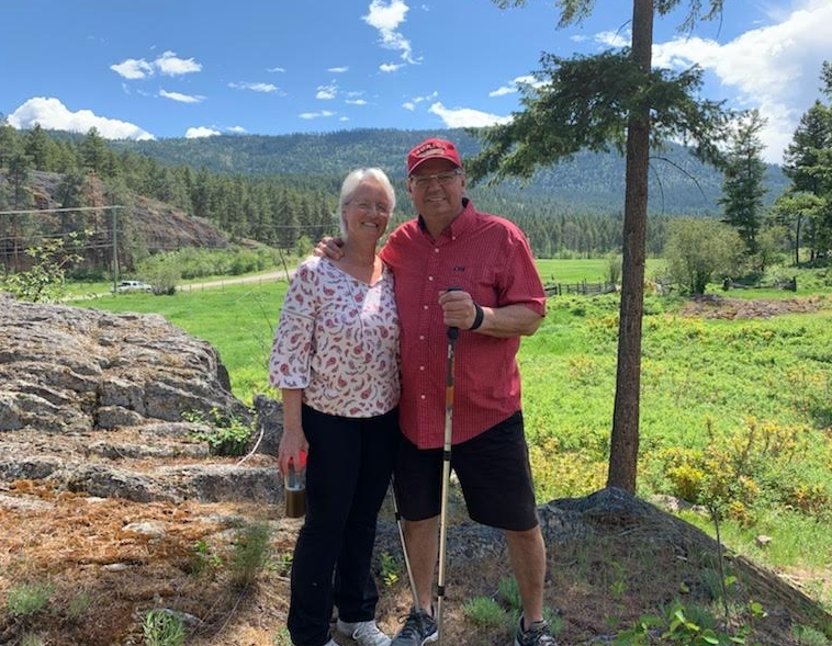 A smiling elderly woman and a middle-aged man stand outdoors on a sunny day, embracing each other. They are surrounded by a green landscape with trees and mountains in the background.  TK Glamping and Wellness in Vernon BC