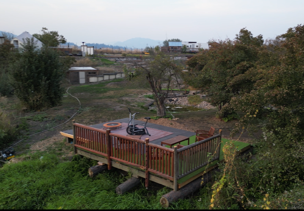 An outside deck next to a babbling brook with available exercise equipment, surrounded by trees and a grassy yard part of a coulee, overlooking open land and distant mountains. TK Glamping and Wellness in Vernon BC.