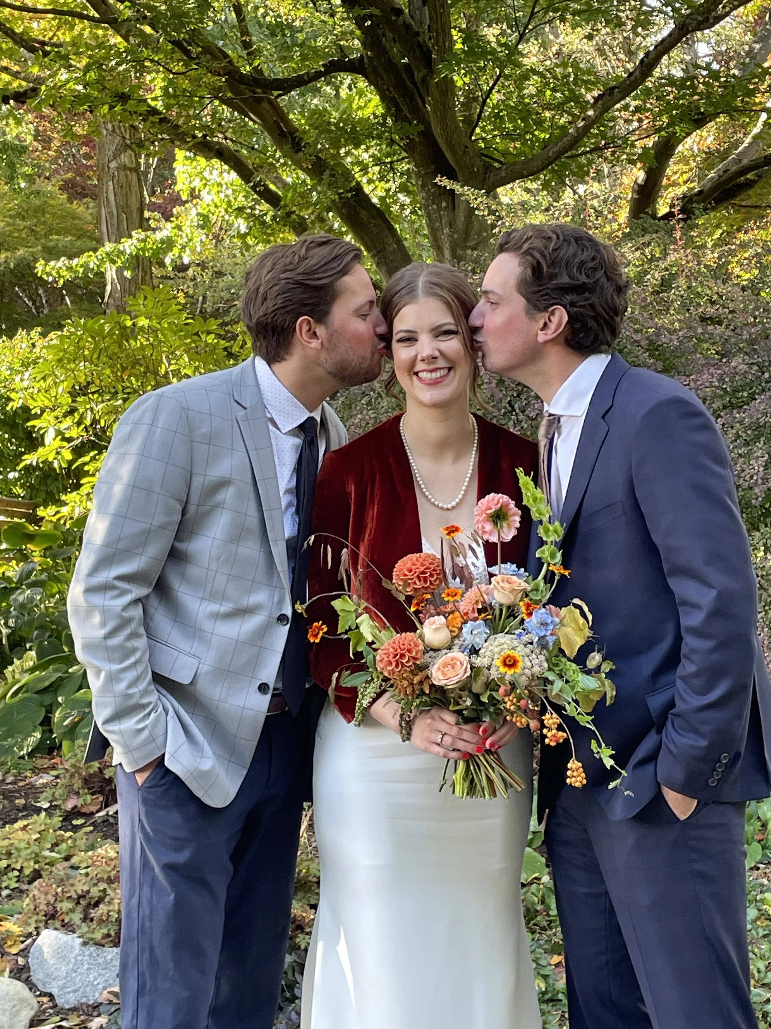 A bride in a white dress and pearl necklace, holding a bouquet of colorful flowers, is flanked by two men in suits, both kissing her on the cheeks in a garden setting with trees and green foliage. TK Glamping and Wellness in Vernon BC.