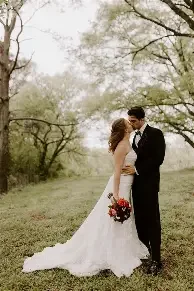 A wedding photo with bride holding flower bouquet wearing a white wedding dress and groom in black suit and bow tie are standing outdoors on grass with trees in the background, embracing each other. TK Glamping and Wellness in Vernon BC.