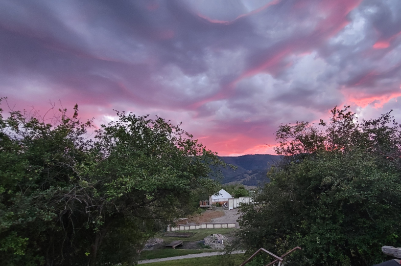 A landscape view of a sunset sky with pink and purple clouds, trees and brushes in the foreground, and mountains in the background. Glamping bell tent in picture seating up on a hill valley view.  TK Glamping and Wellness in Vernon BC.