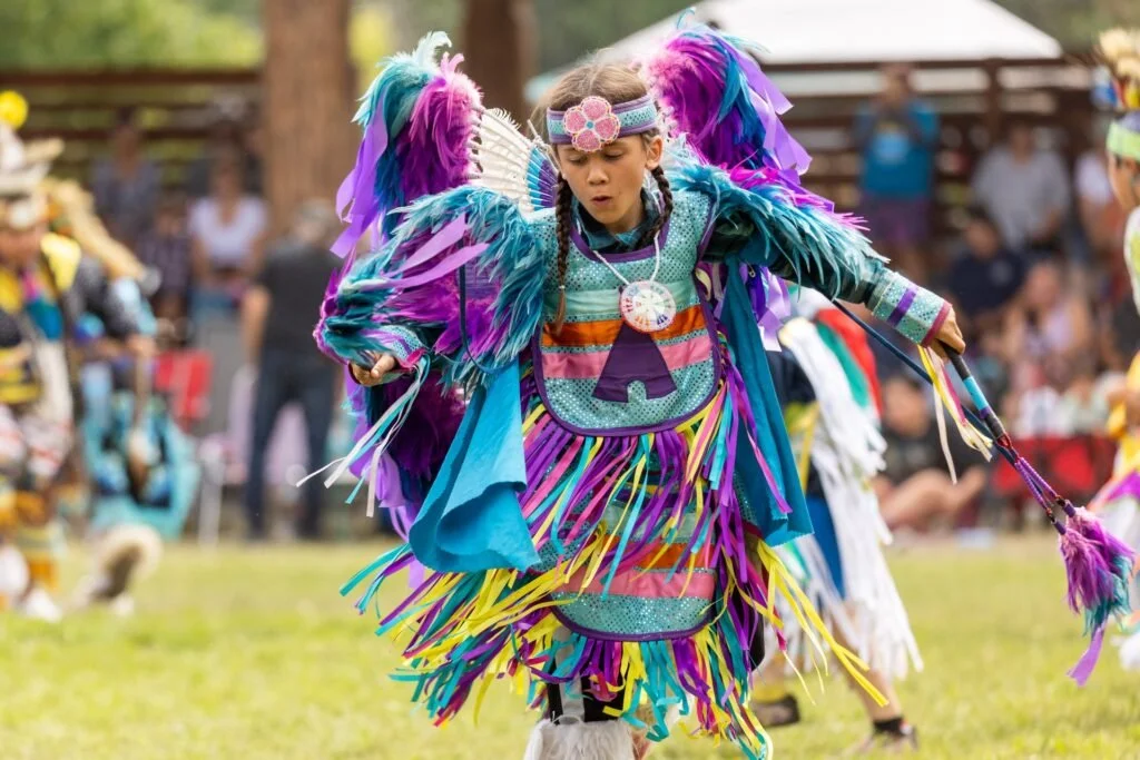 A young indigenous boy wearing a colorful traditional First Nations dance costume with feathers, beads, and fringe, participating in a cultural event or powwow with a crowd in the background.  TK Glamping and Wellness in Vernon BC.