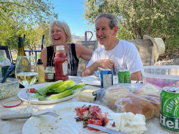 Two people, an elderly man and woman, are sitting at a table outdoors, laughing and enjoying a meal. The table has various food items, drinks, and condiments, with trees and a clear sky in the background. TK Glamping and Wellness in Vernon BC.
