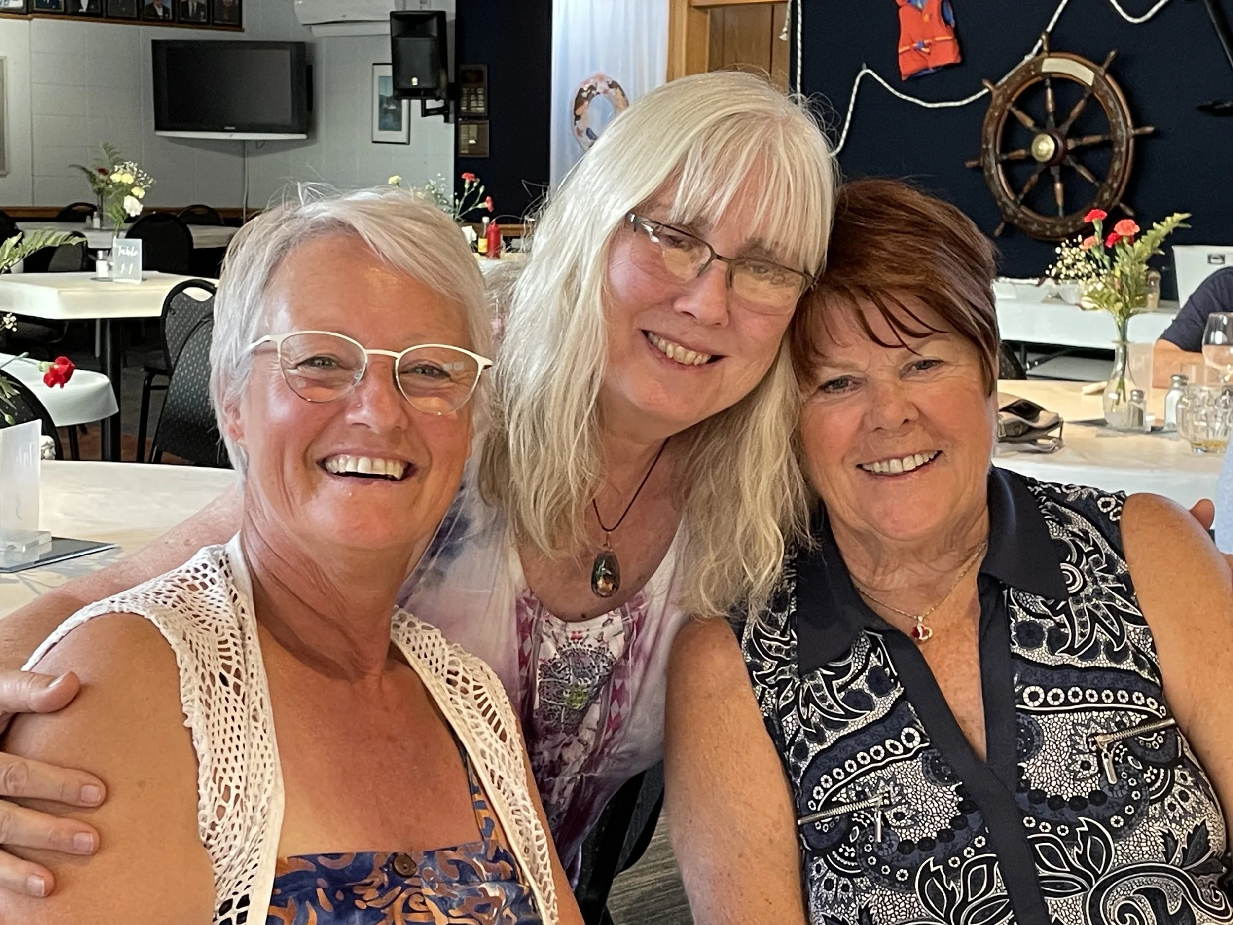 Three women smiling, sitting close together indoors, with table, flowers, and nautical-themed decor in the background. Good friends enjoying each others company and imagining their time together while glamping.  TK Glamping and Wellness in Vernon BC.