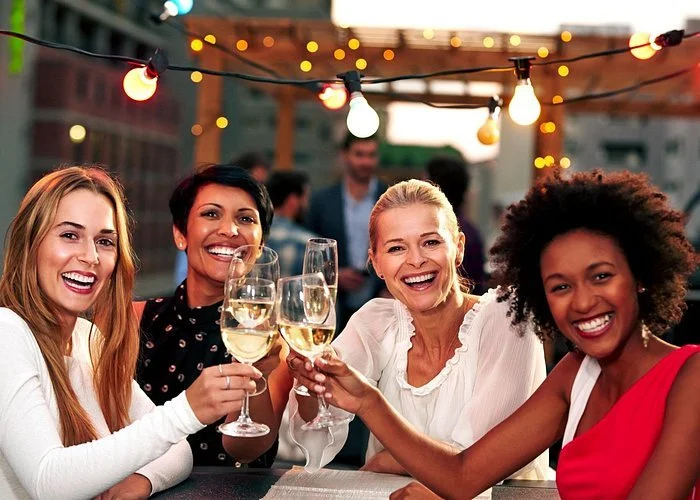 Four women smiling and toasting with wine glasses at an outdoor party during evening, decorated with string lights. Enjoying an Okanagan Lake wine tour in and around Vernon and Kelowna. TK Glamping and Wellness in Vernon BC.