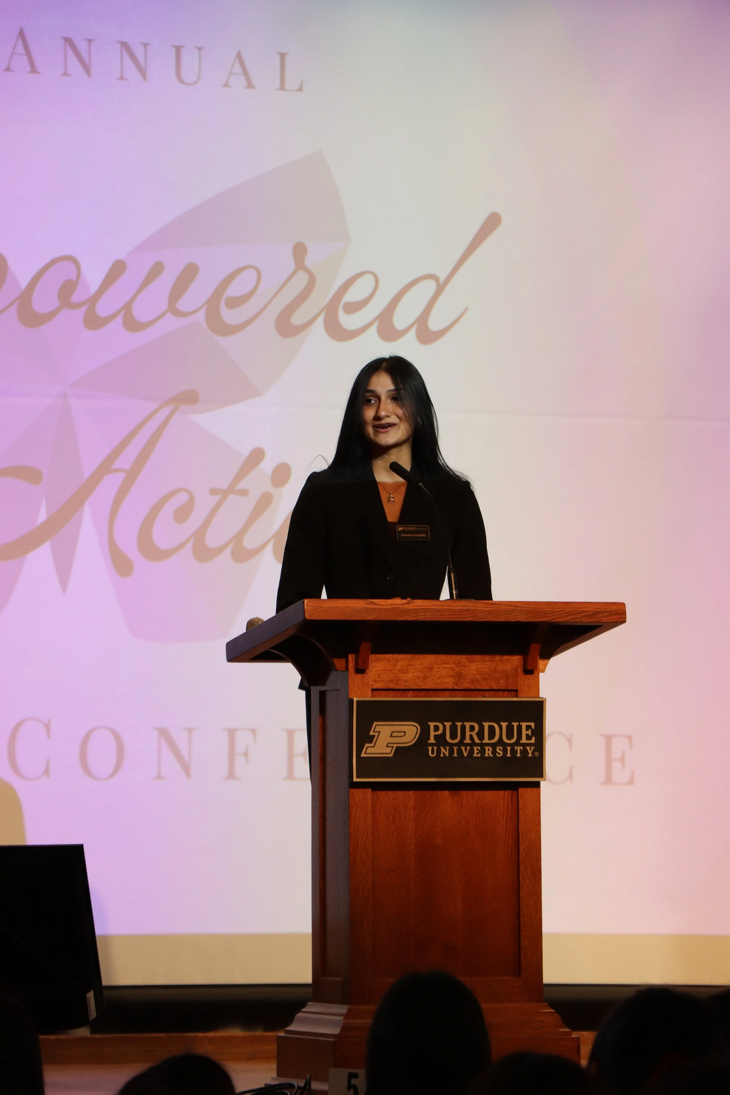 A young woman giving a speech at a podium during an event at Purdue University, with a large screen behind her displaying the words 'Powered Action.'