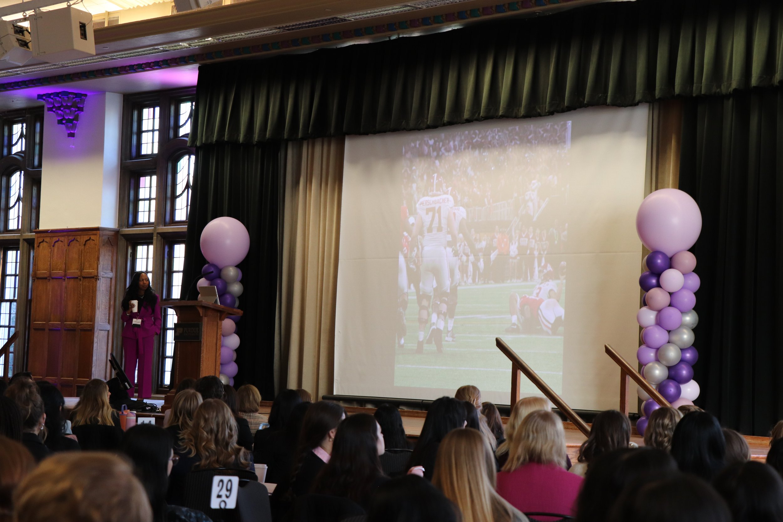 A woman in a magenta suit speaking at a podium on a stage in a large auditorium or conference room, with an audience seated facing the stage. There are large balloon columns on either side of a projection screen displaying a football game.
