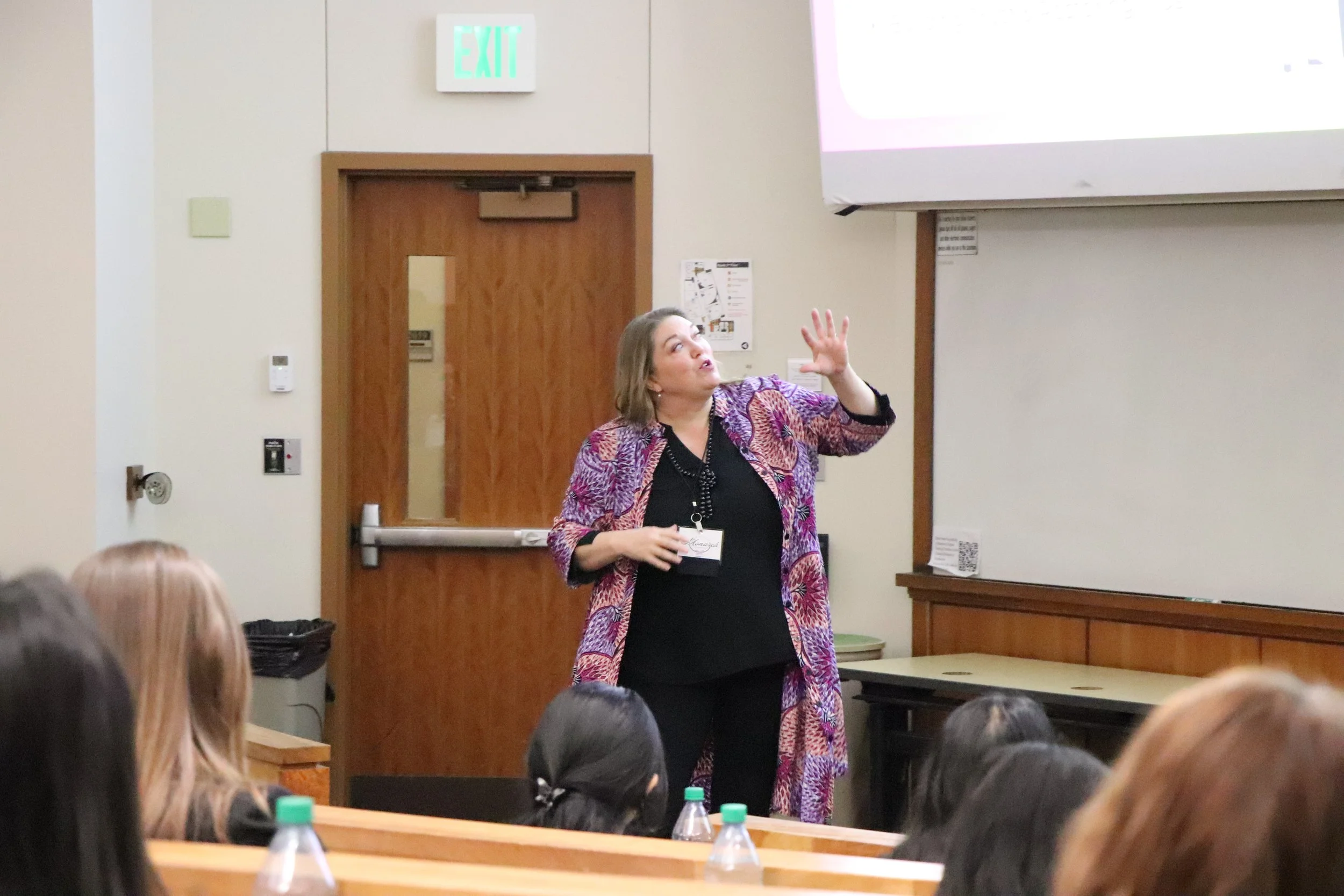 A woman standing in front of a classroom giving a presentation, with students seated in the foreground and an exit sign in the background.