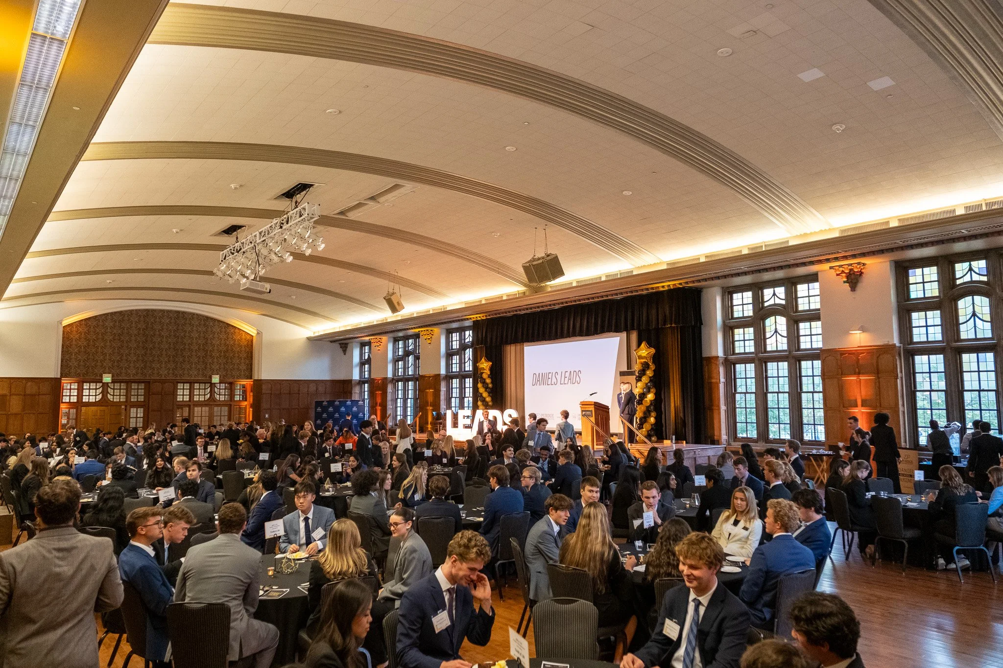 Large indoor event with many people seated at round tables, a stage with a large screen and people standing, decorated with balloons, and balloons spelling out "LEADS" at the front.